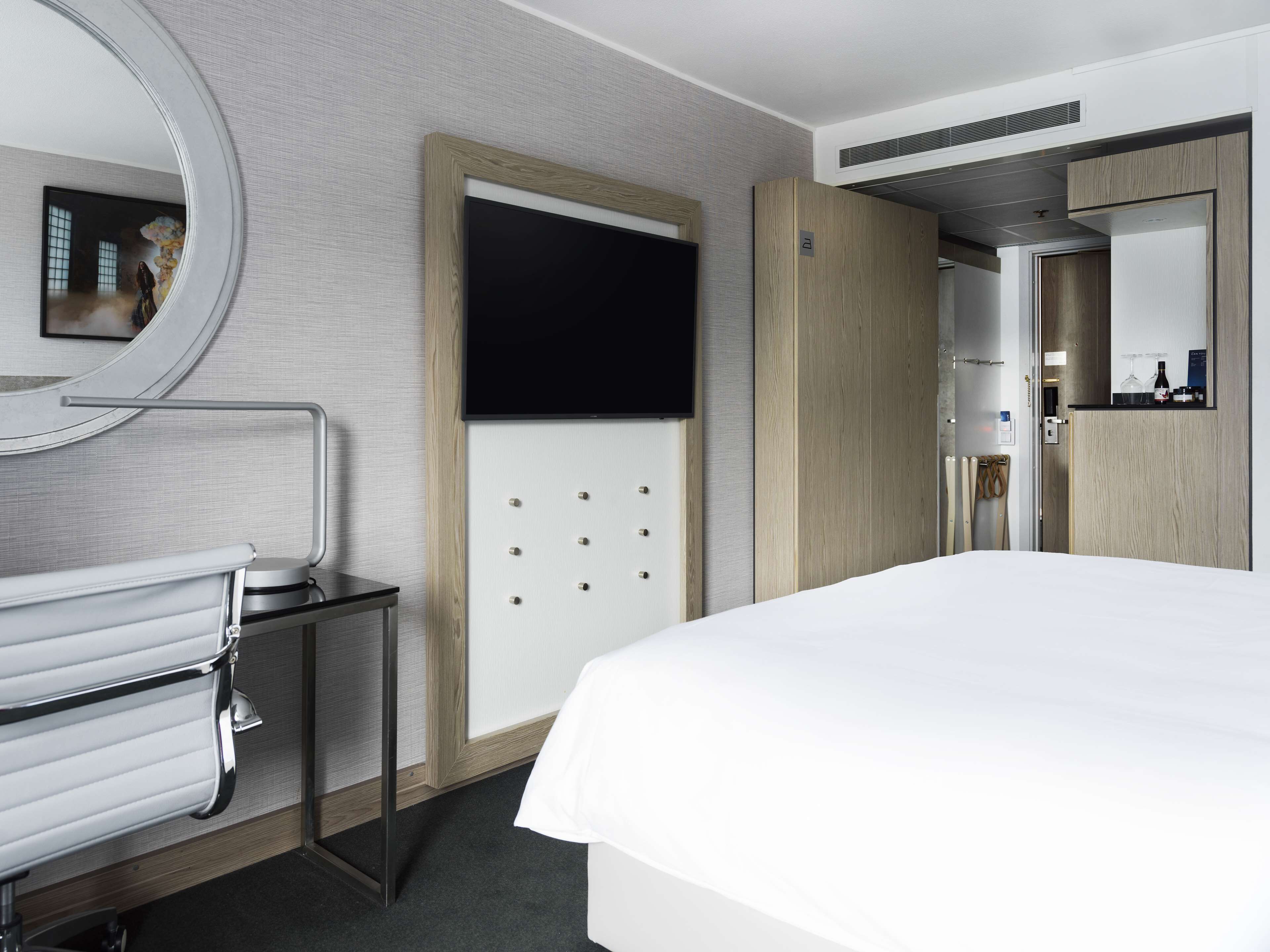 A modern hotel room with a large bed, a round mirror, a small table with a chair, a wooden wardrobe, and a bathroom with a toilet and sink.
