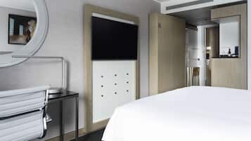 A modern hotel room with a large bed, a round mirror, a small table with a chair, a wooden wardrobe, and a bathroom with a toilet and sink.