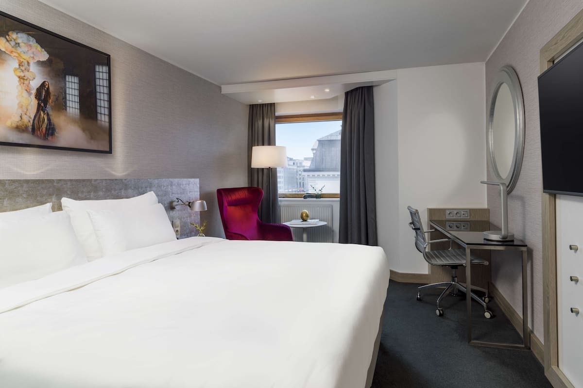 Une chambre d’hôtel avec un grand lit, un fauteuil rouge, un bureau avec un miroir et un téléviseur fixé au mur.