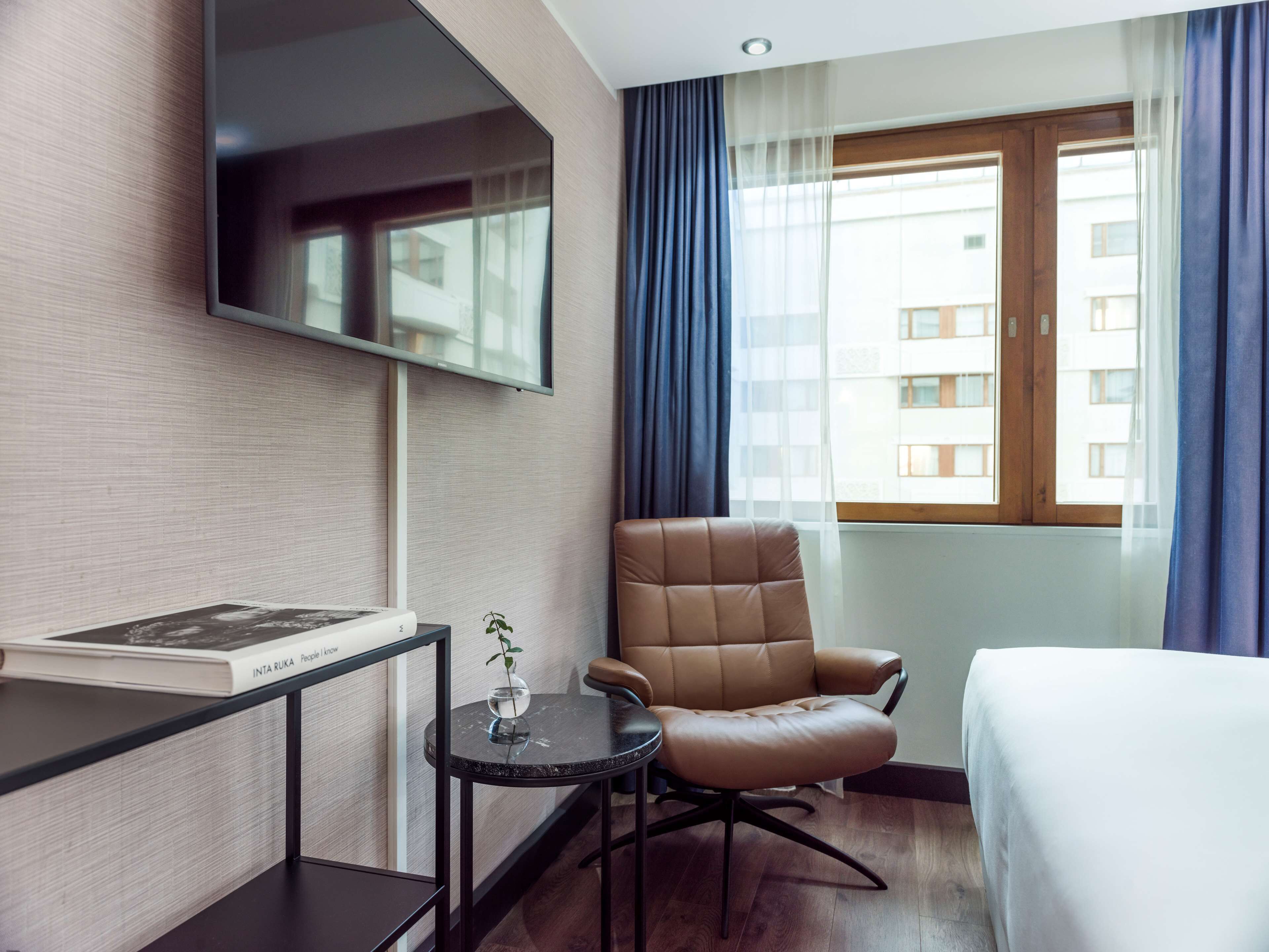 Una habitación de hotel con una cama, una silla, una mesita con un libro, un televisor montado en la pared y una ventana con cortinas.