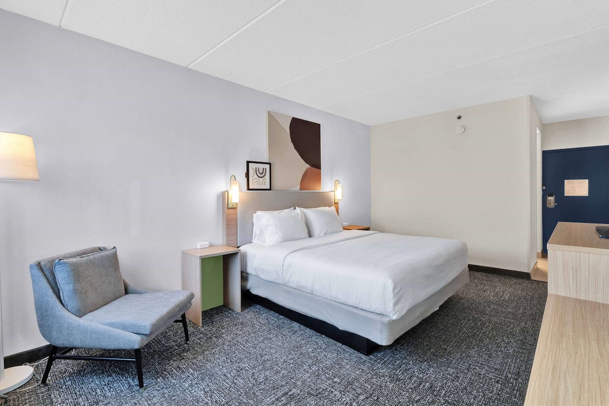 Een hotelkamer met een bed, een stoel, een nachtkastje en een kunstwerk aan de muur.