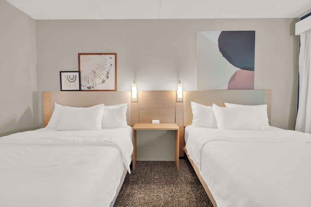 Een hotelkamer met twee bedden, een houten hoofdbord, een nachtkastje met een lamp en een kunstwerk aan de muur.