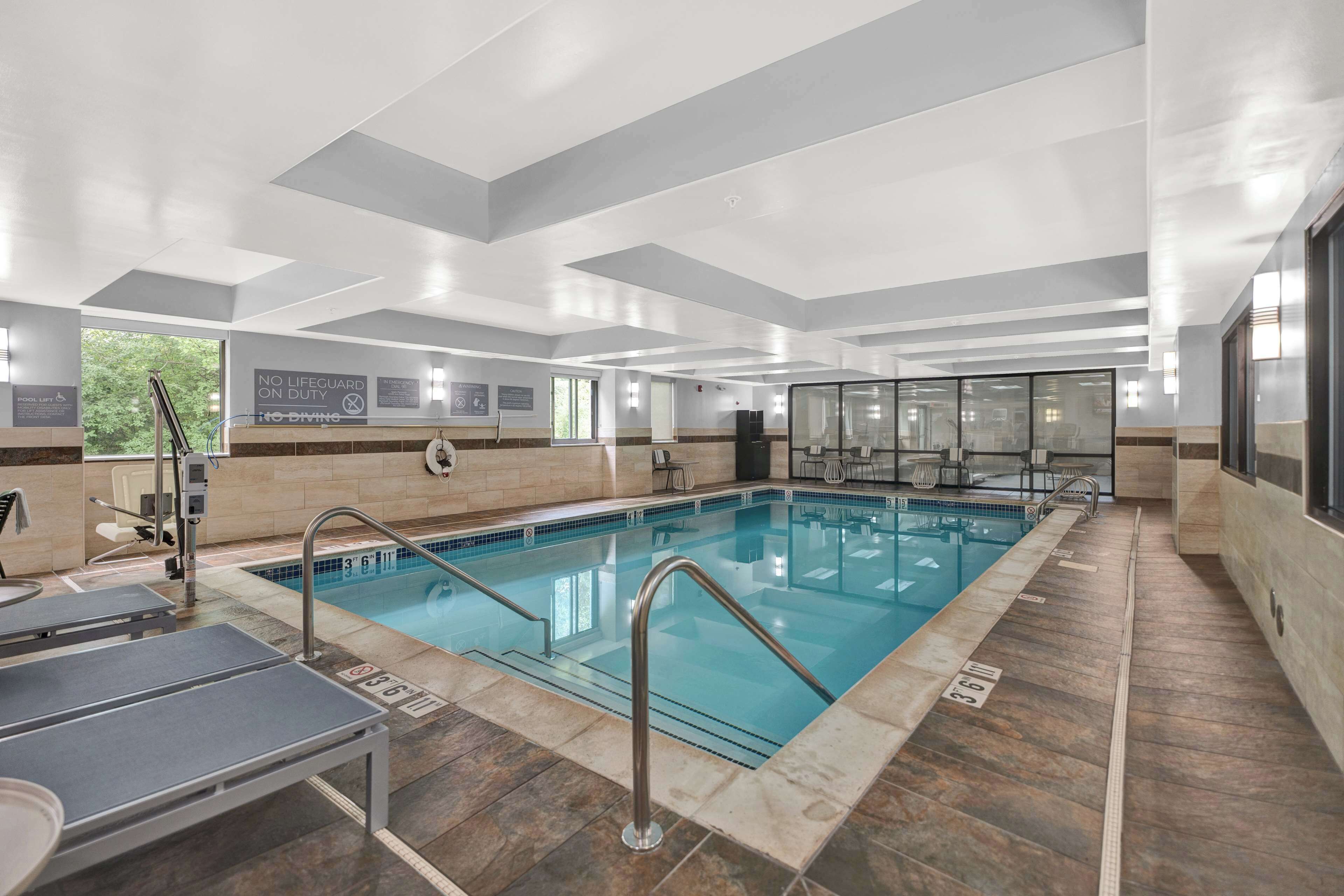 En moderne indendørs swimmingpool med tydelig skiltning, en redningsstation og et solbelyst område med siddepladser.