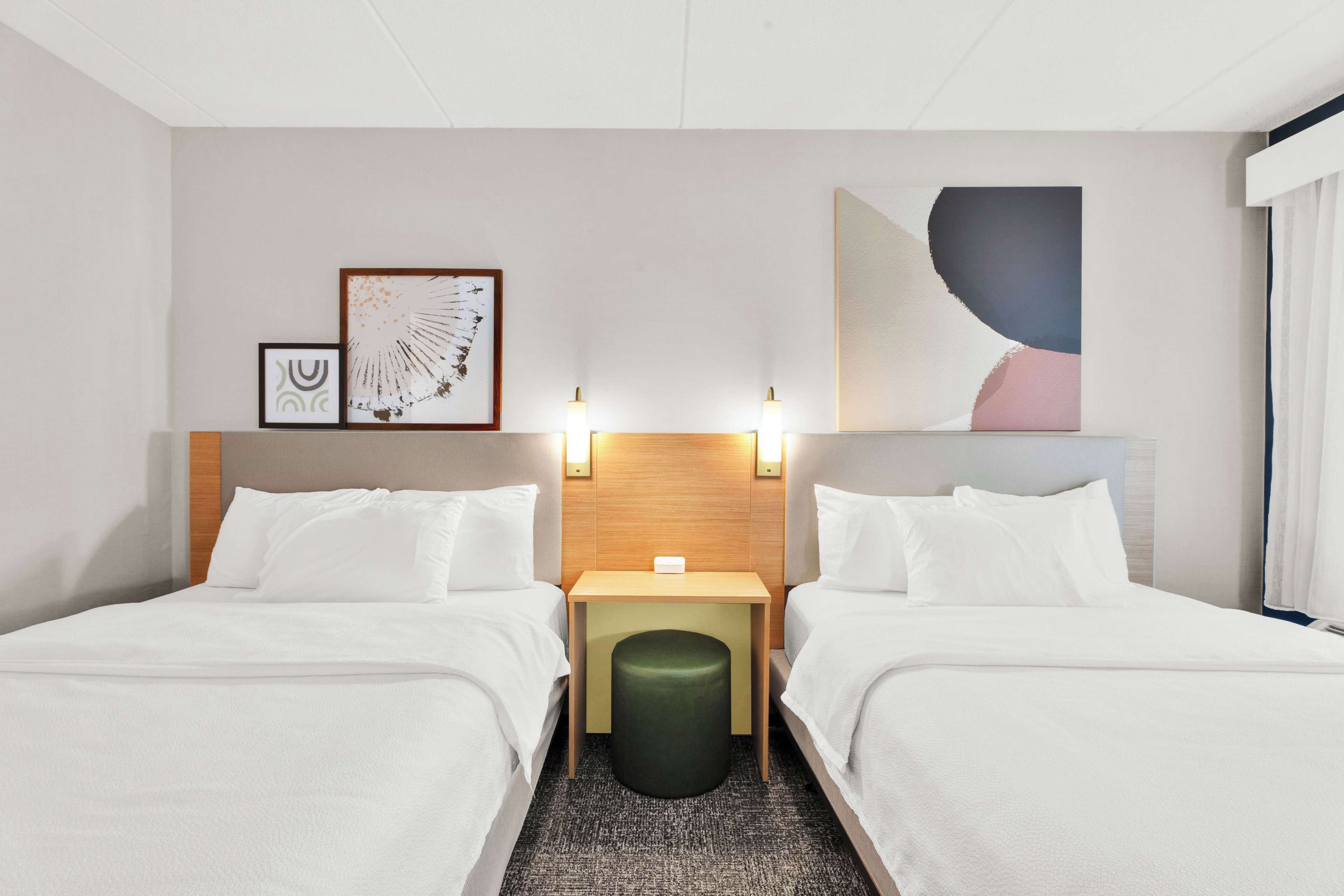 A hotel room with two beds, a small table, a green ottoman, and wall art.