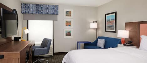 A hotel room with a bed, a desk with a TV, a chair, a lamp, and framed pictures on the wall.