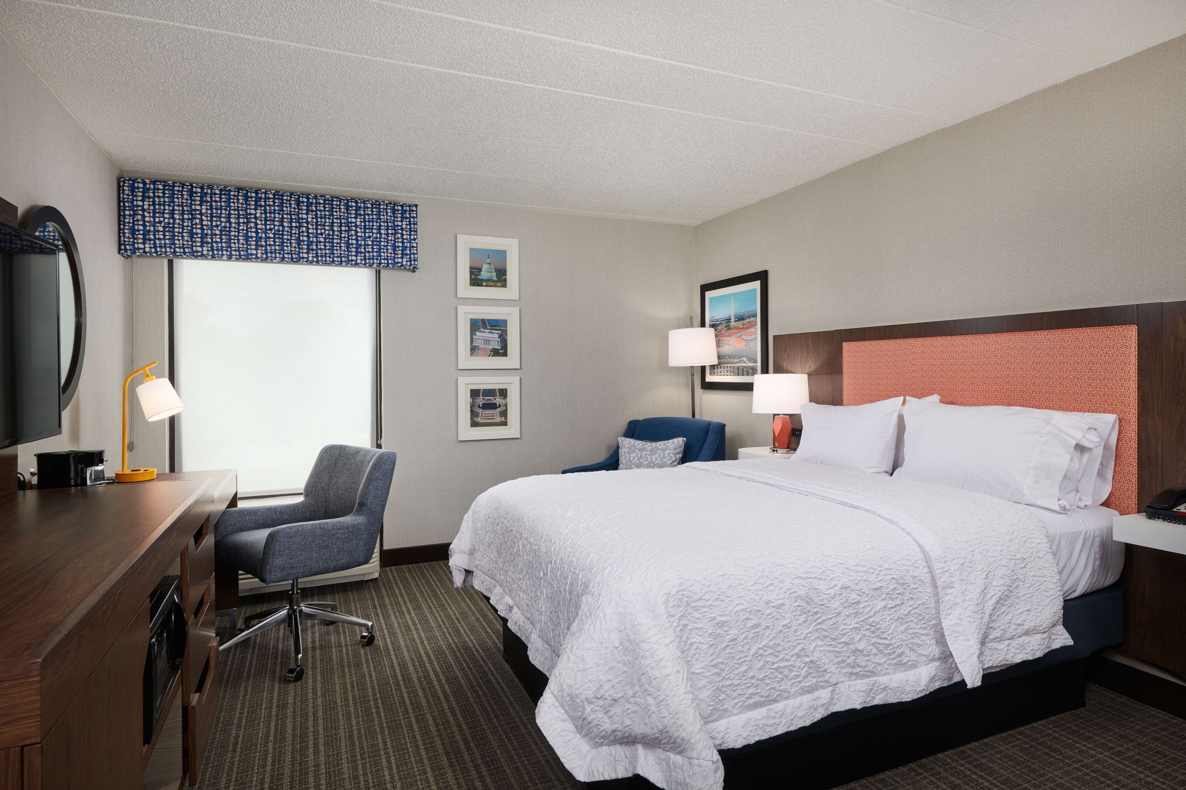 A hotel room with a large bed, a desk with a chair, a television, and framed pictures on the wall.