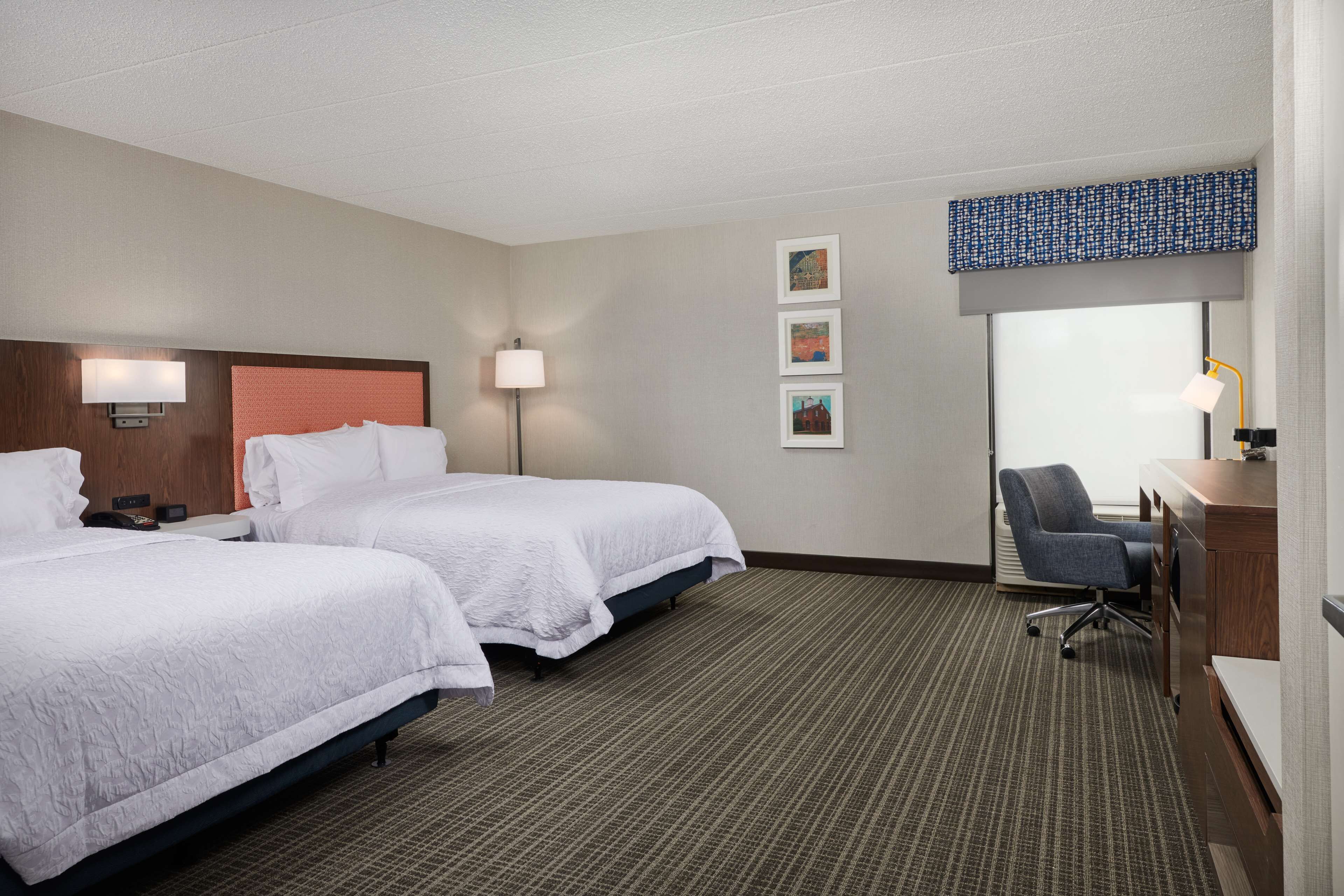 A hotel room with two beds, a desk, a chair, a lamp, and artwork on the wall.