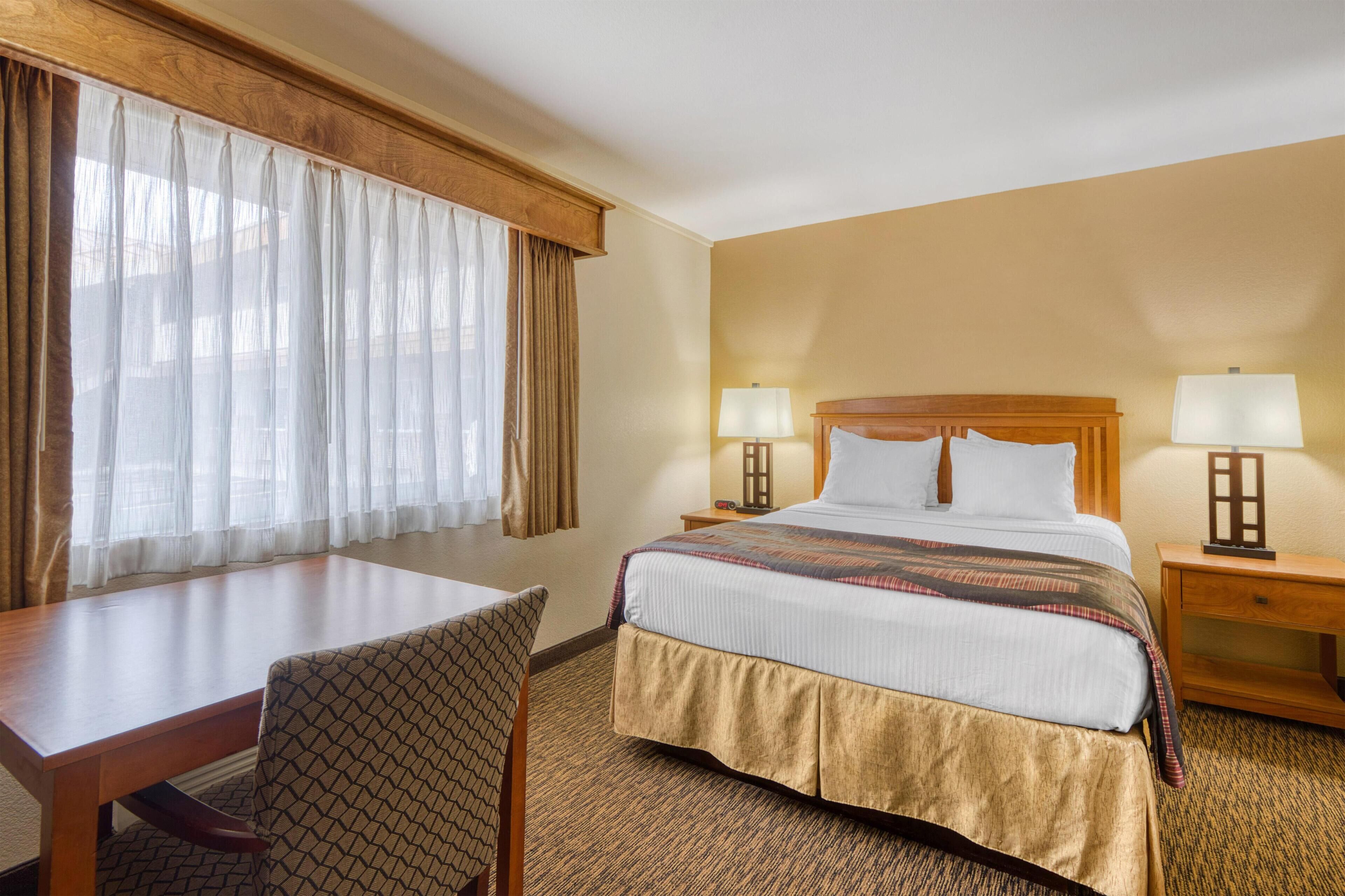 Camera d'albergo con un letto, due comodini con lampade, una scrivania con sedia e una finestra con tende.