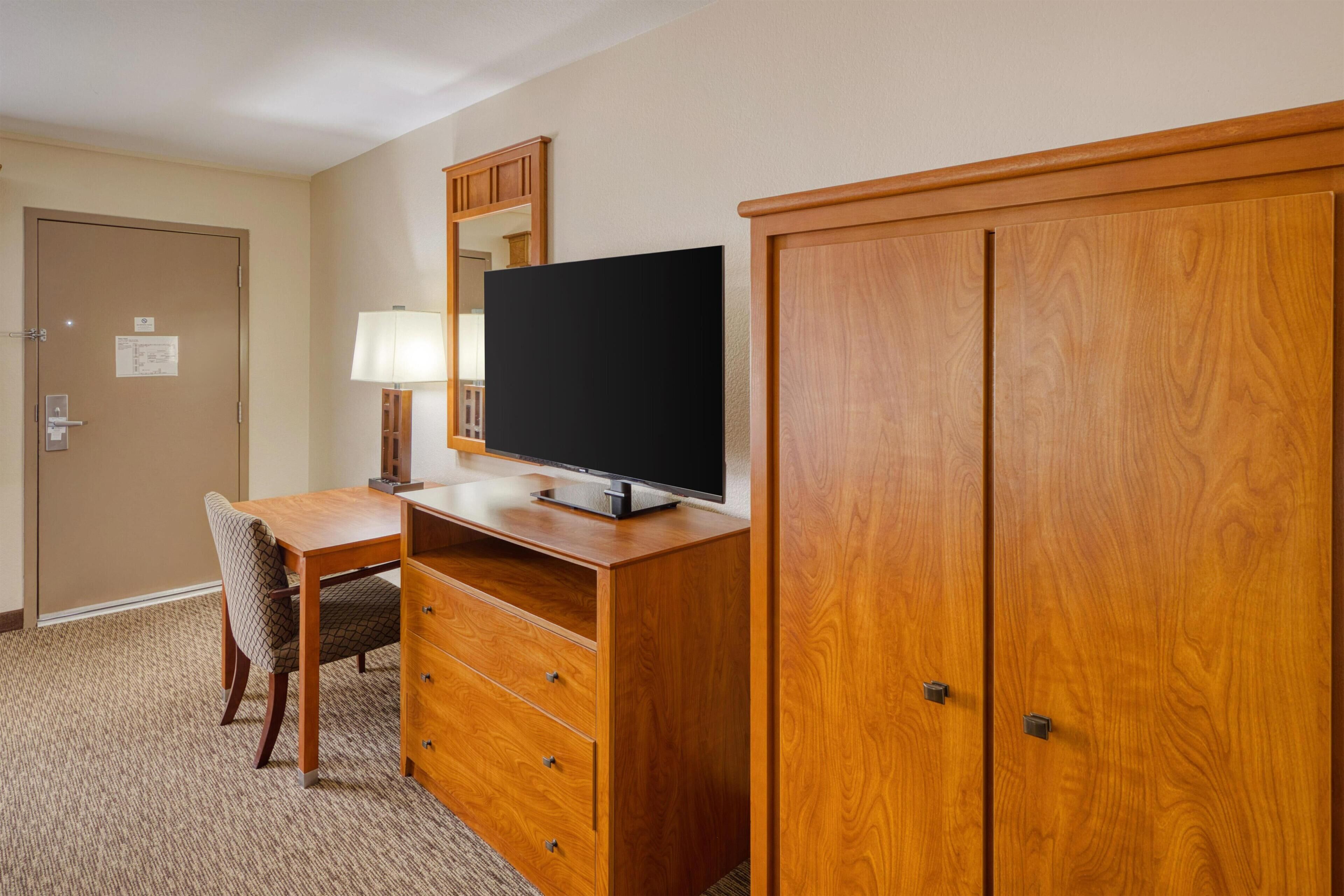 Ein Hotelzimmer mit Schreibtisch aus Holz, einem Fernseher, einem Sessel, einer Lampe und einem Kleiderschrank.
