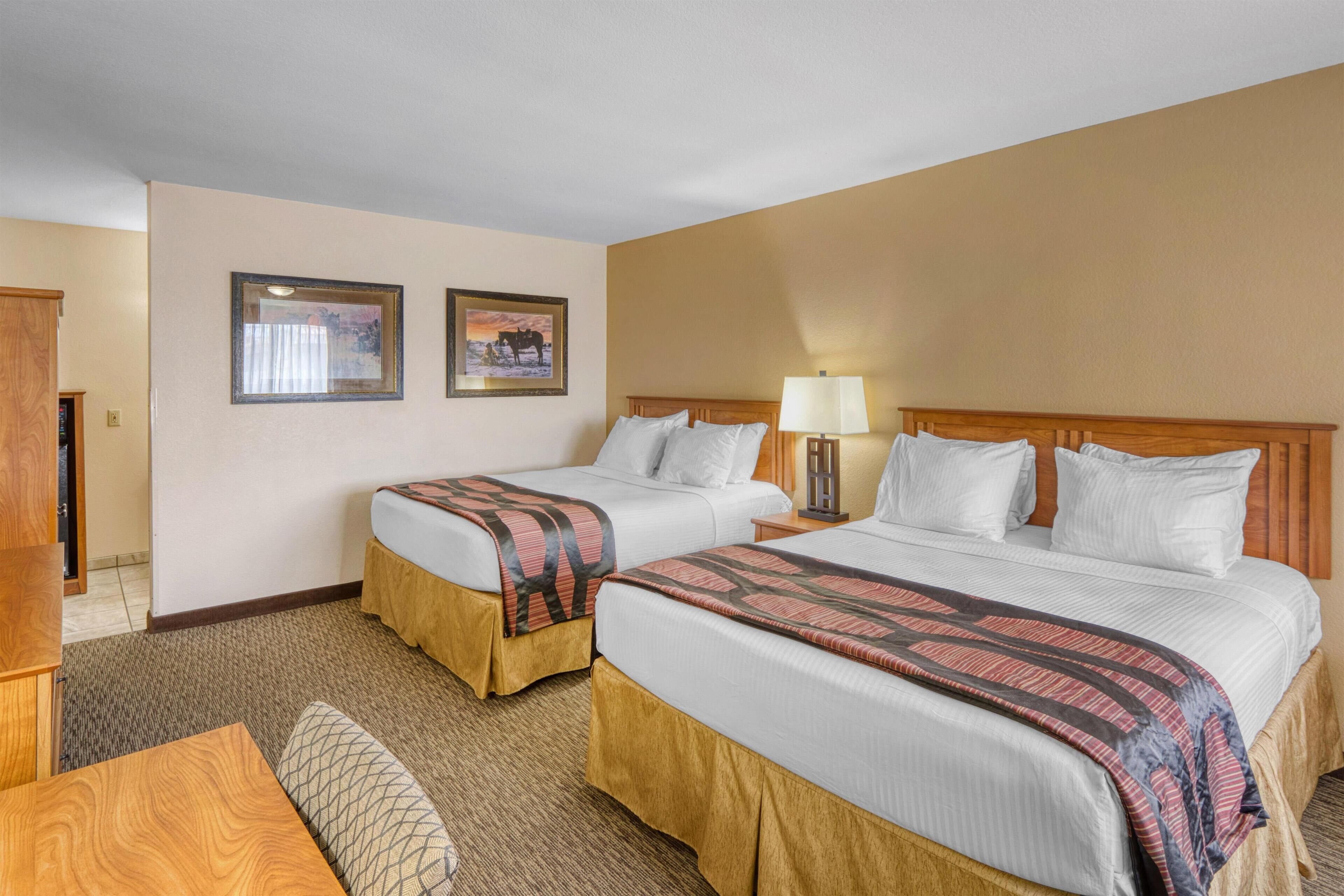 A hotel room with two beds, a desk, a lamp, and two framed pictures on the wall.