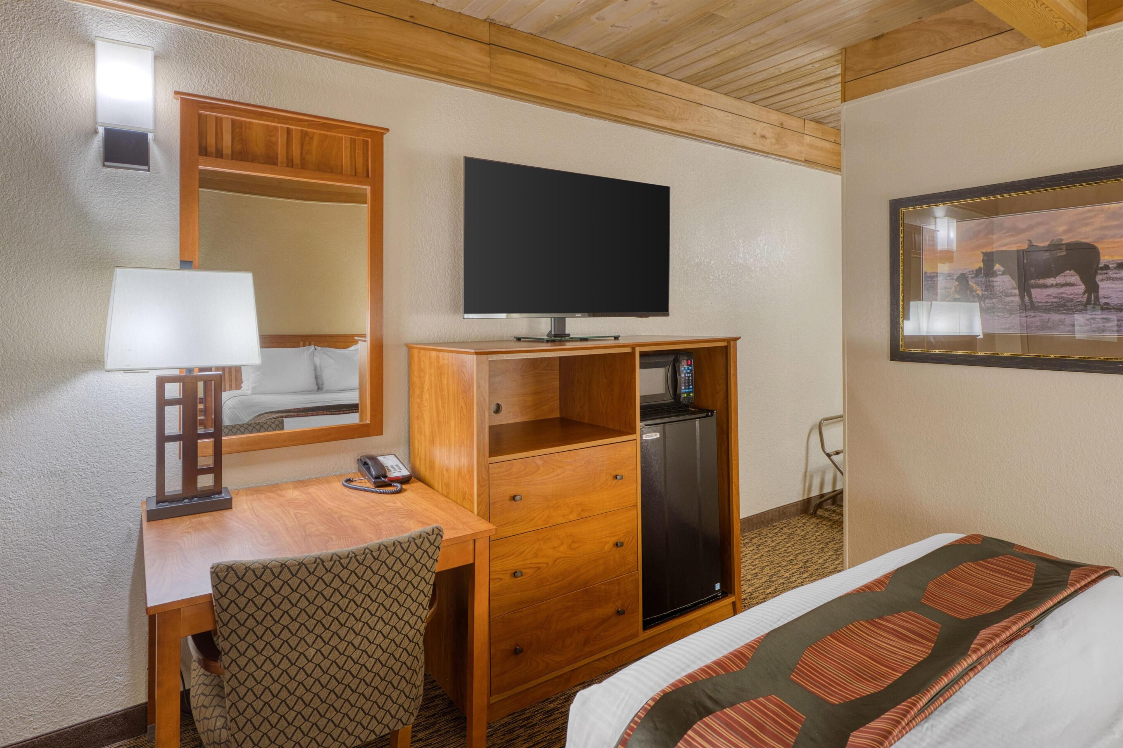 Habitación de hotel con un escritorio de madera, televisión, refrigerador, cama con ropa de cama estampada, una silla e imagen enmarcadas en la pared.