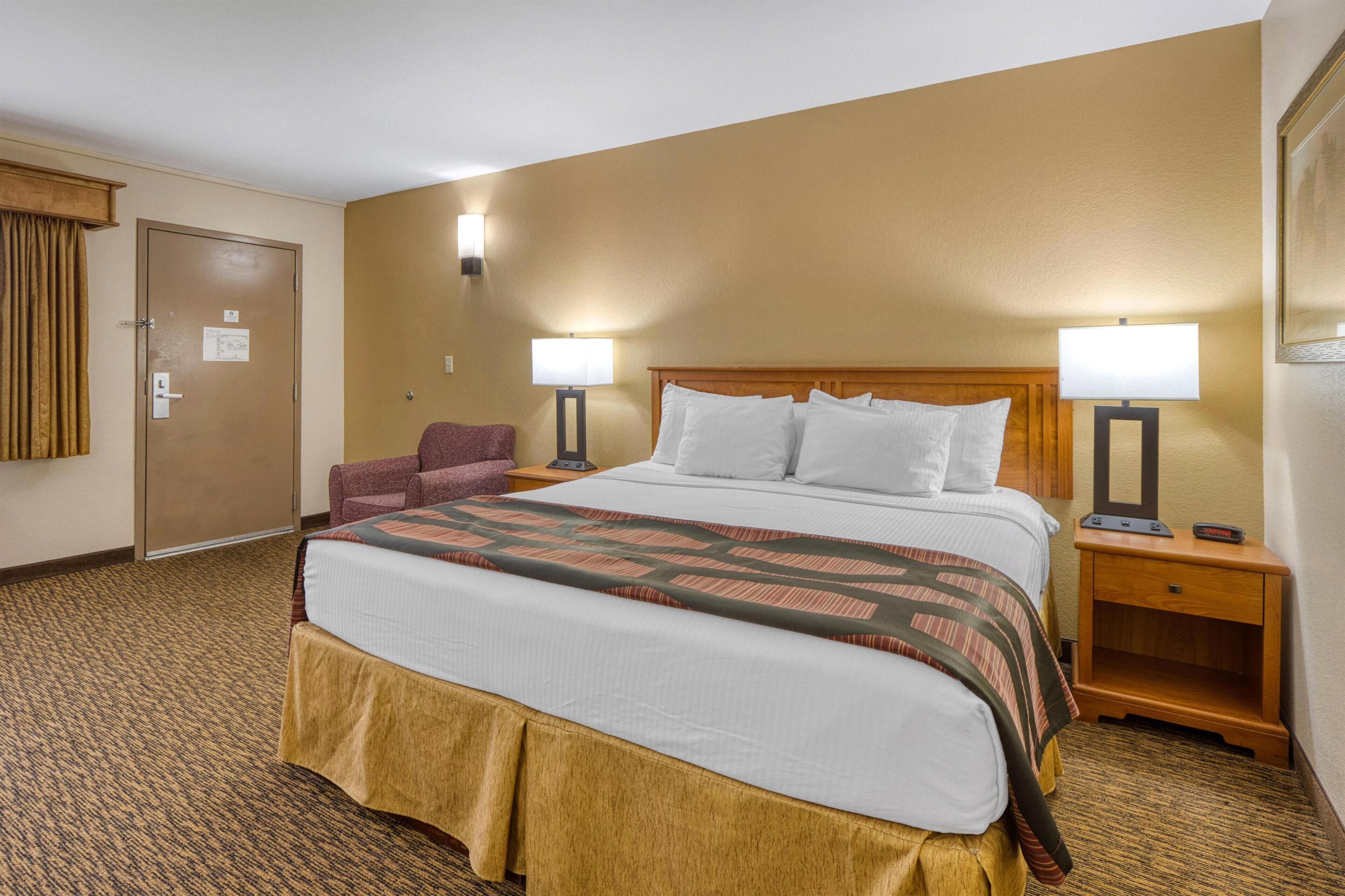 A hotel room with a large bed, two bedside lamps, a chair, a wooden nightstand, and a door.