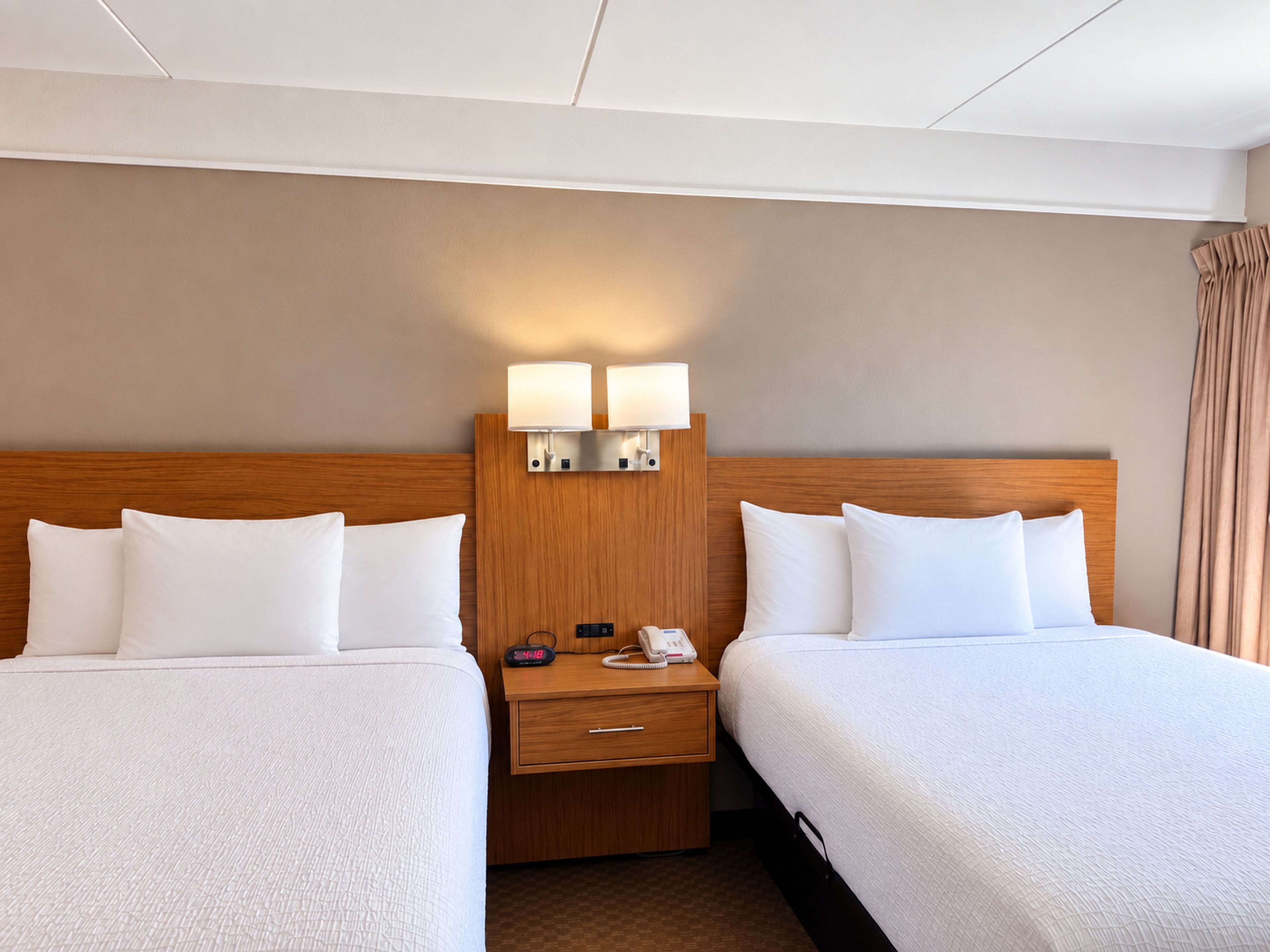 Una camera d'albergo con due letti, una testiera in legno, comodini con lampade e un tavolino con una sveglia.