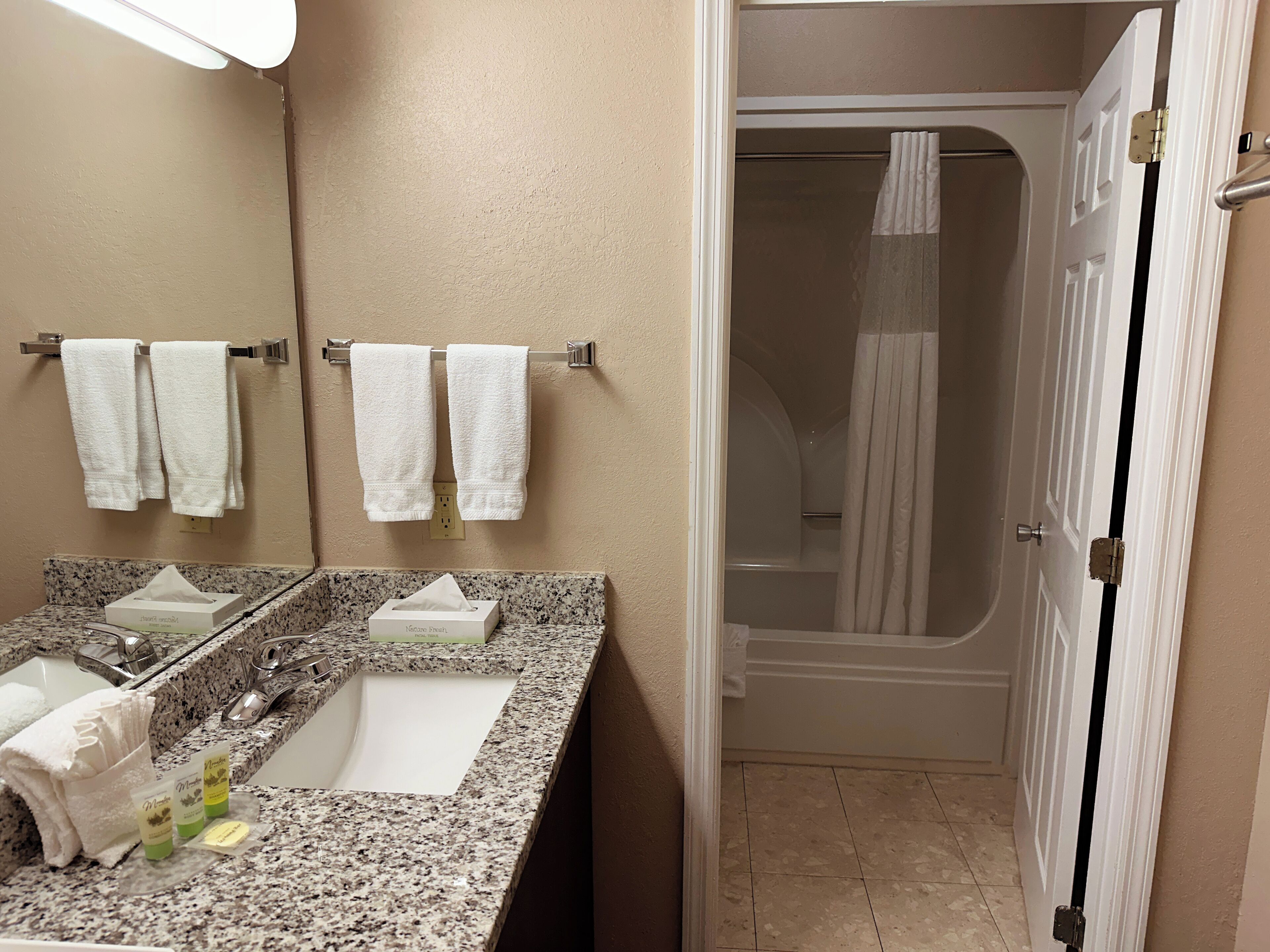 A bathroom with a granite countertop, two sinks, a mirror, and a shower area.