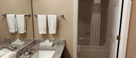 A bathroom with a granite countertop, two sinks, a mirror, and a shower area.