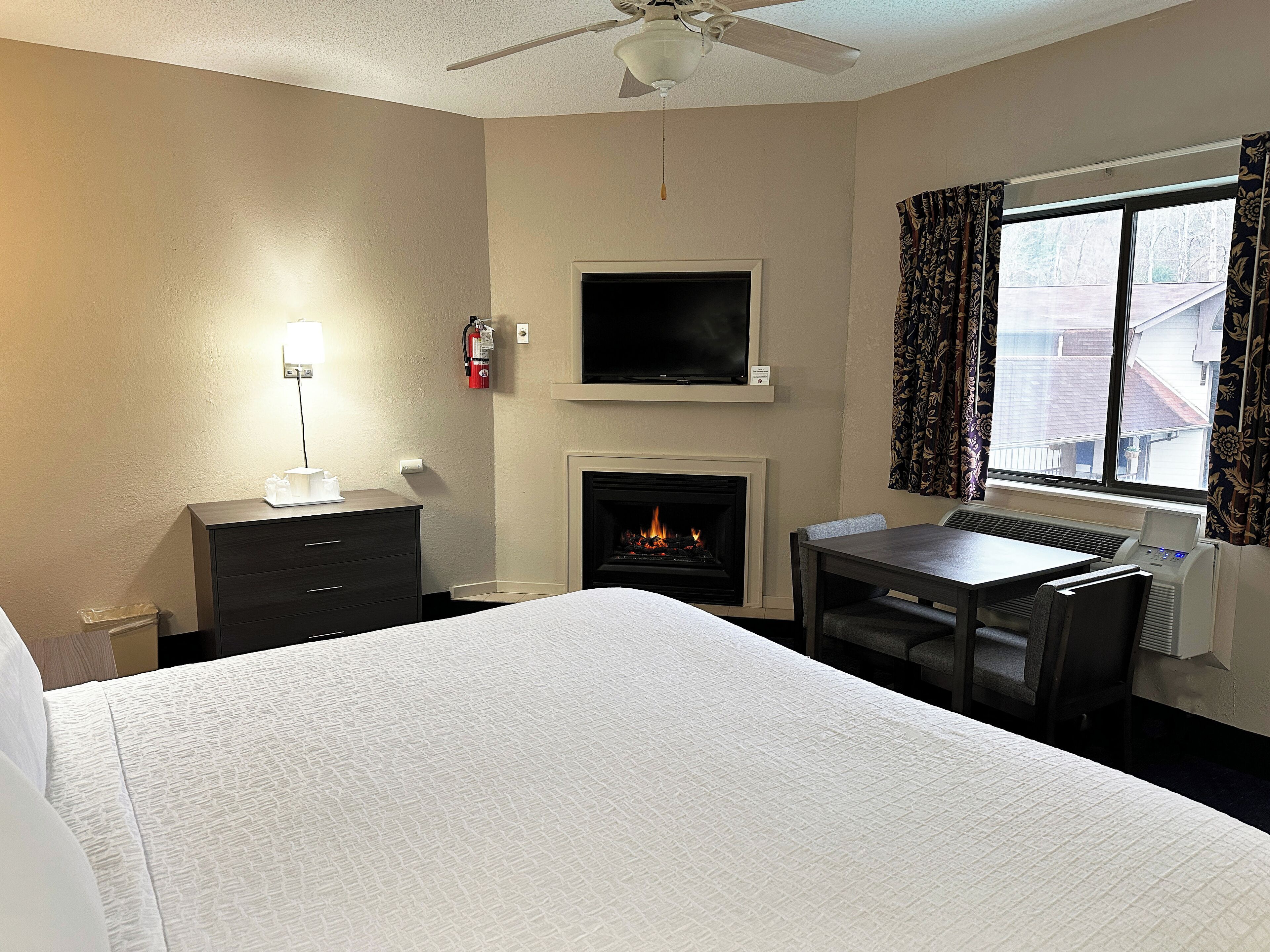 A hotel room with a bed, a desk, a fireplace, a TV, and a window with curtains.