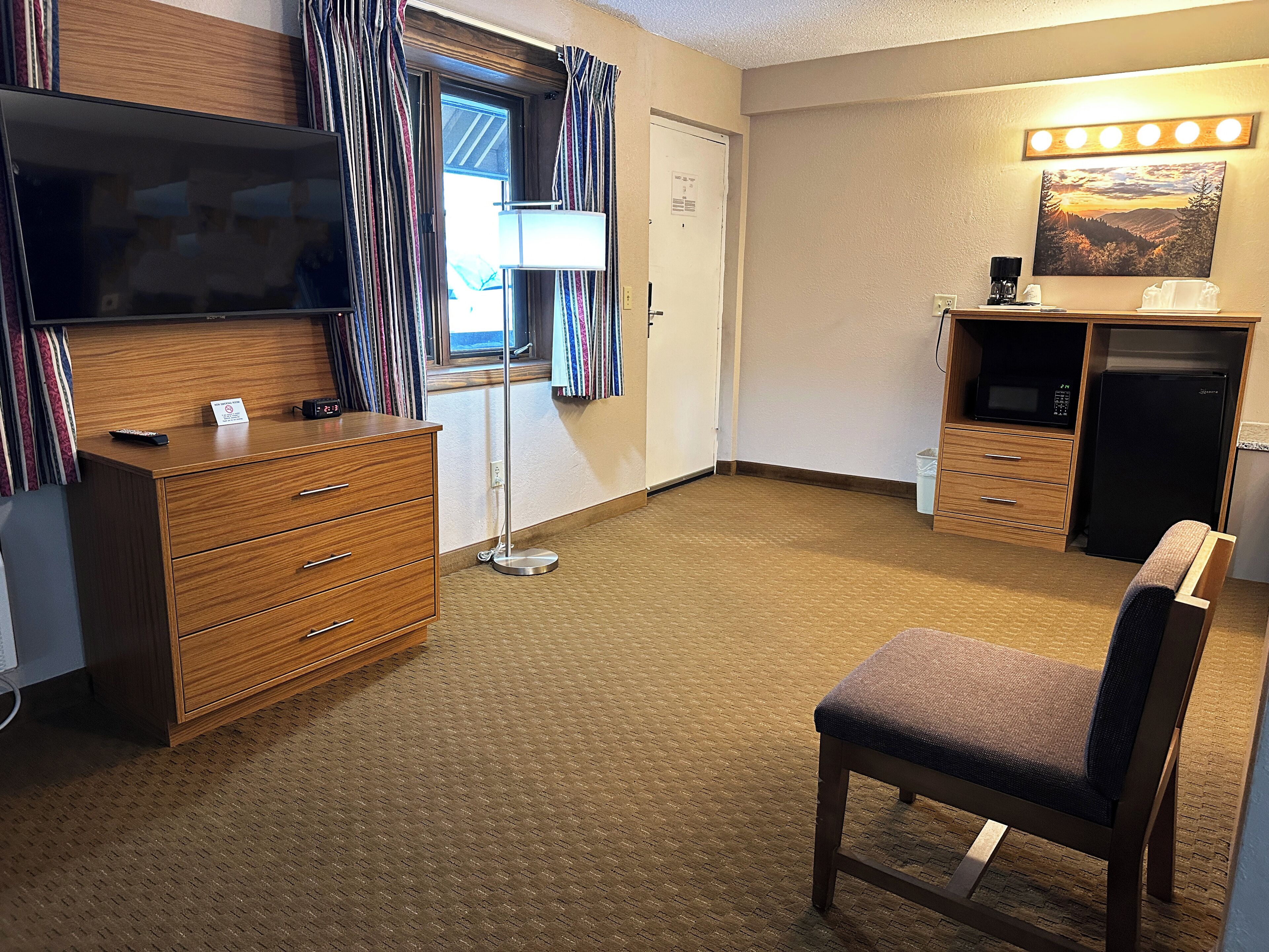 Une chambre d’hôtel avec une commode en bois, un téléviseur, un bureau avec une lampe, une chaise, un four à micro-ondes, un réfrigérateur et un tableau au mur.