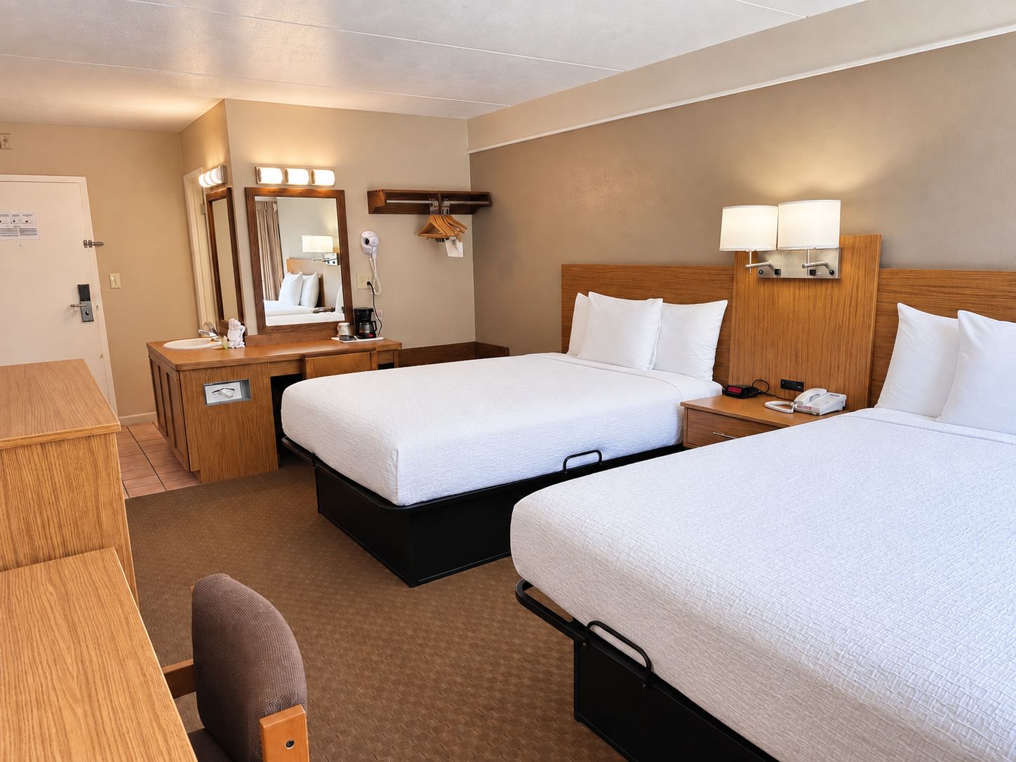 A hotel room with two beds, a desk, a mirror, and a bedside table with lamps.
