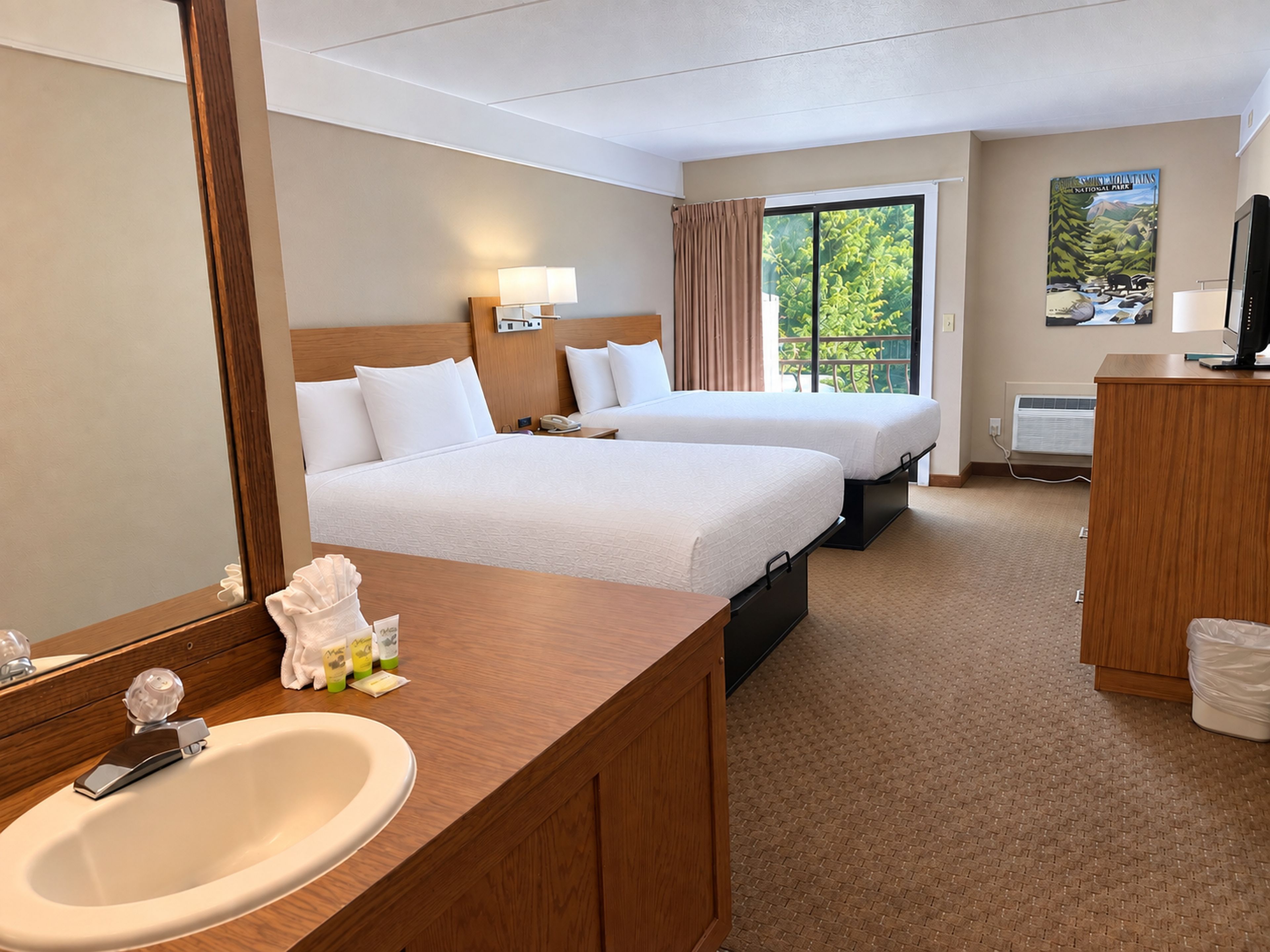 Habitación de hotel con dos camas, un espejo grande, un aparador de madera, un lavamanos y una ventana con vista a follaje.