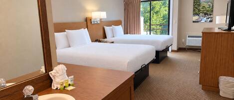 Habitación de hotel con dos camas, un espejo grande, un aparador de madera, un lavamanos y una ventana con vista a follaje.
