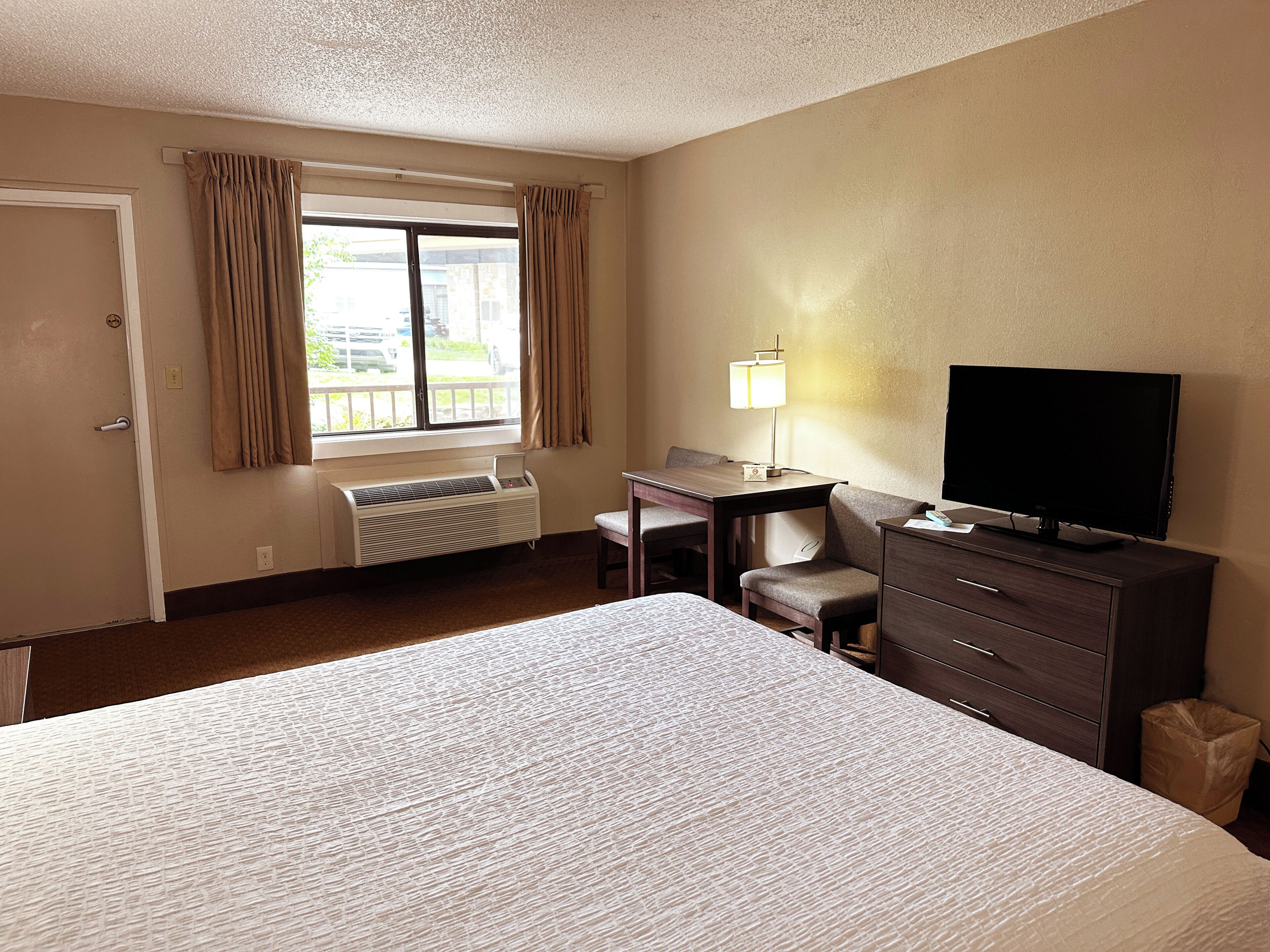 Een hotelkamer met een bed, een bureau met stoel, een televisie, een radiator en een raam met gordijnen.