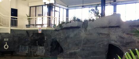 An indoor swimming pool with a rocky backdrop, a balcony with railings, and a ceiling with exposed beams and lighting fixtures.