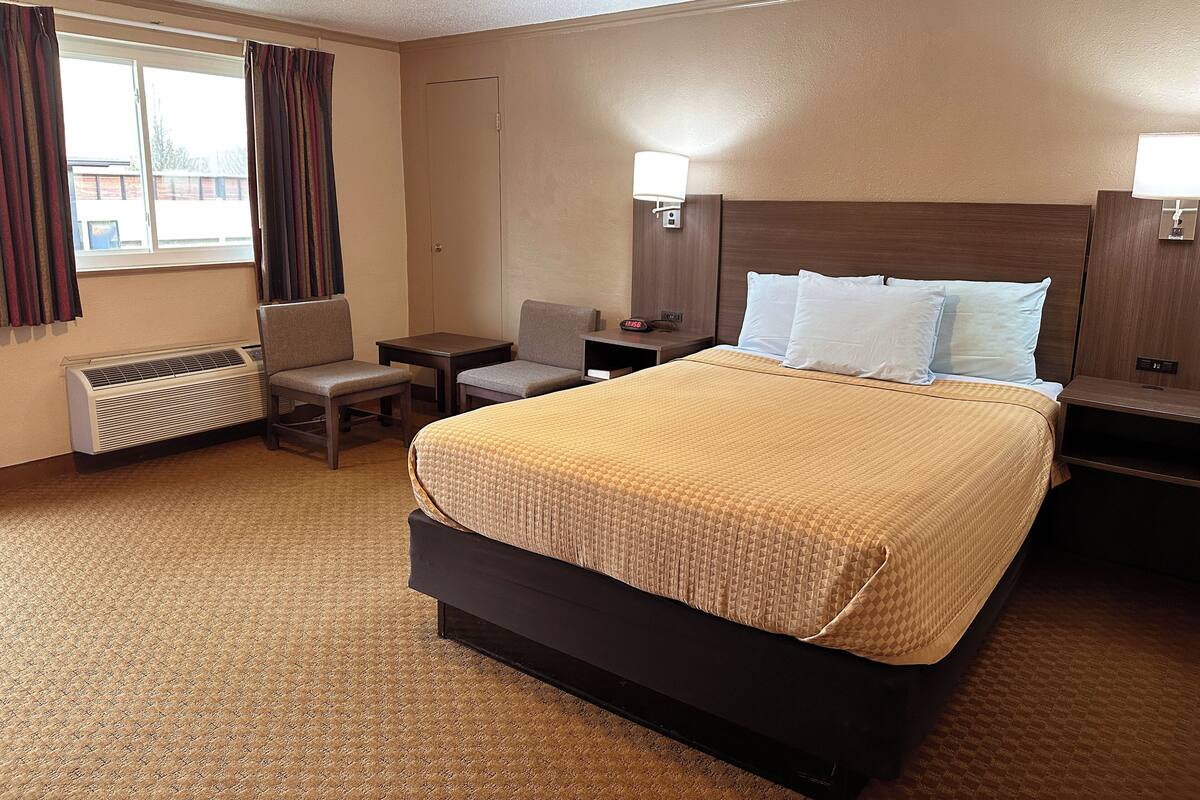 Une chambre d’hôtel avec un grand lit, un bureau, deux chaises, un radiateur et une fenêtre avec des rideaux.