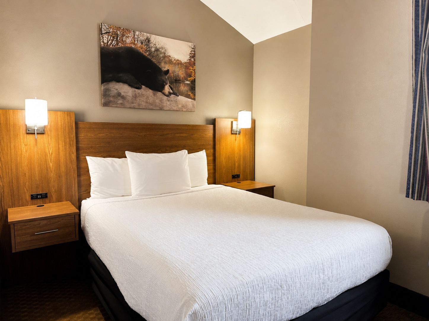 Quarto de hotel com cama grande, duas mesas de cabeceira, cabeceira de madeira e um quadro de urso fixado na parede.