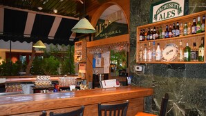 A bar area with wooden bar counter, chairs, and a shelf with bottles.
