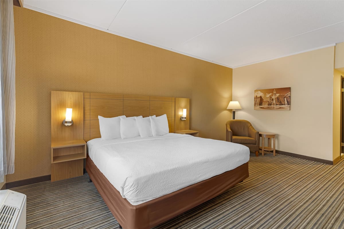 Een hotelkamer met een groot bed, een houten hoofdbord, twee nachtkastjes met lampen, een kleine stoel en een schilderij aan de muur.