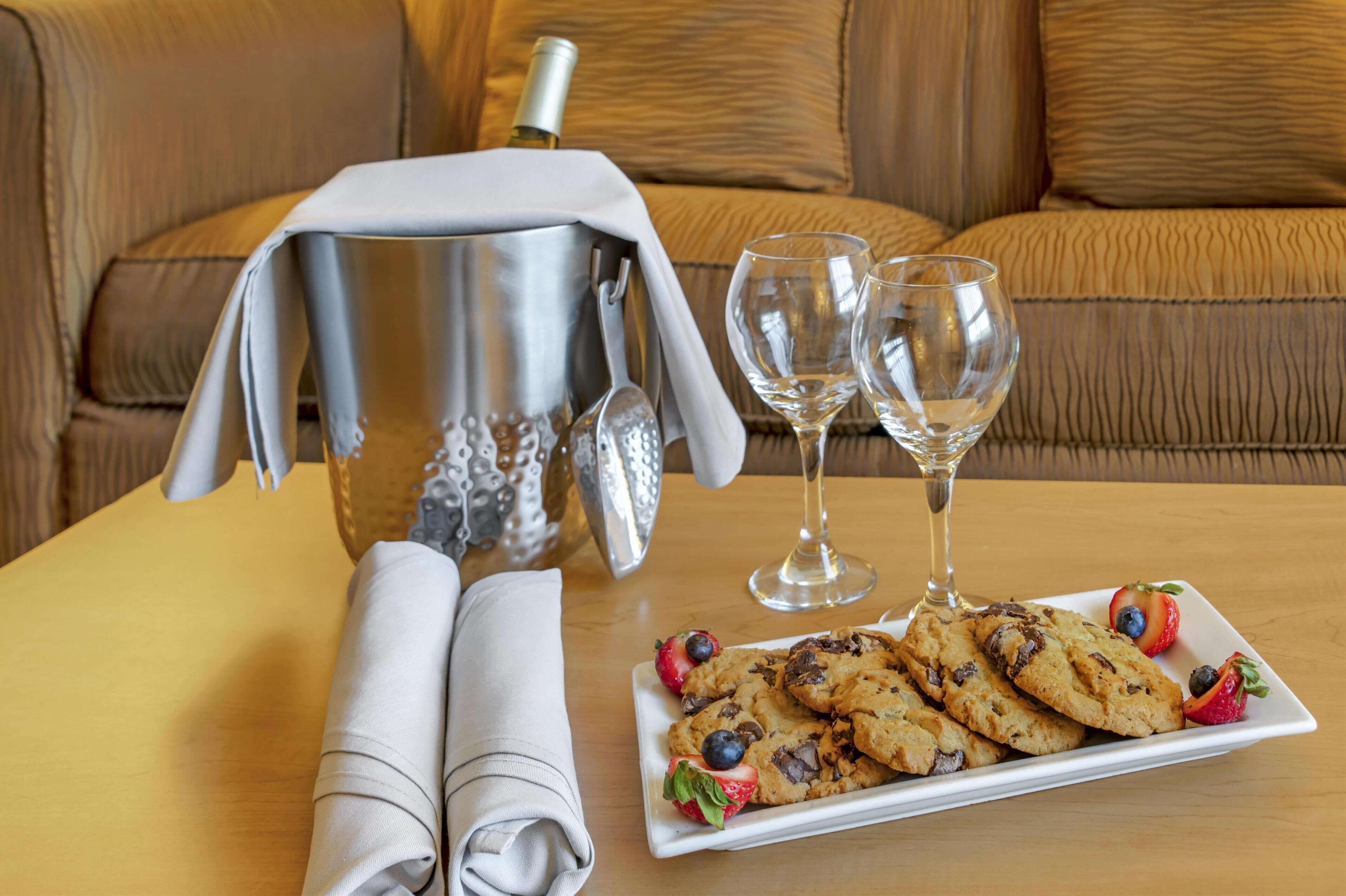 Una mesa ordenada con un plato de galletas, dos copas de vino, una botella y una jarrón plateado.