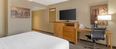 A hotel room with a bed, a desk with a chair, a flat-screen TV, a mirror, and artwork on the walls.