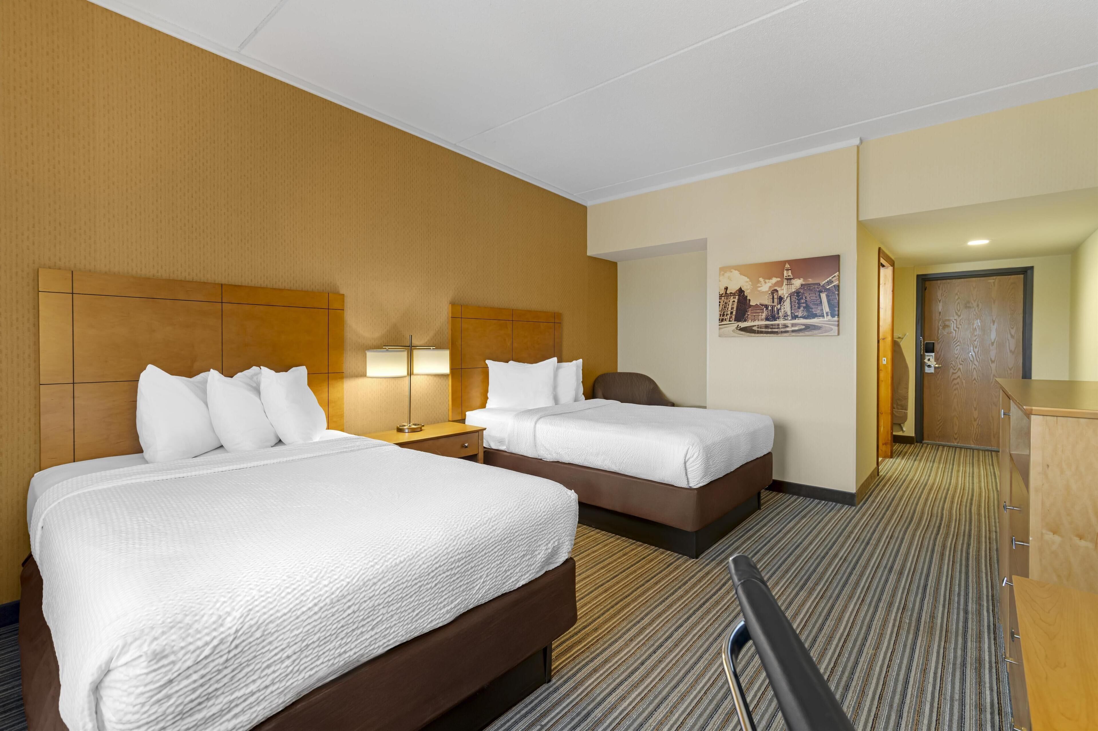 Quarto de hotel com duas camas, mesa de cabeceira com luminárias, cabeceira de madeira, quadro na parede e cômoda de madeira.