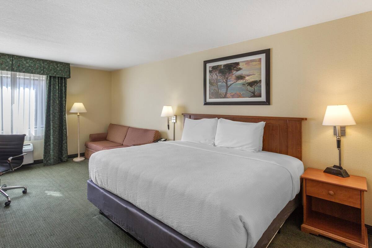 A hotel room with a large bed, two bedside lamps, a desk with a chair, and a painting on the wall.
