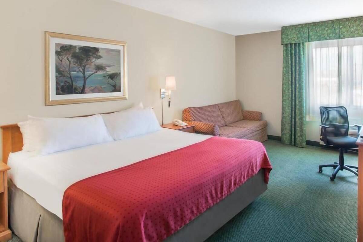 A hotel room with a bed, a desk, a chair, a lamp, a painting, and a window with curtains.