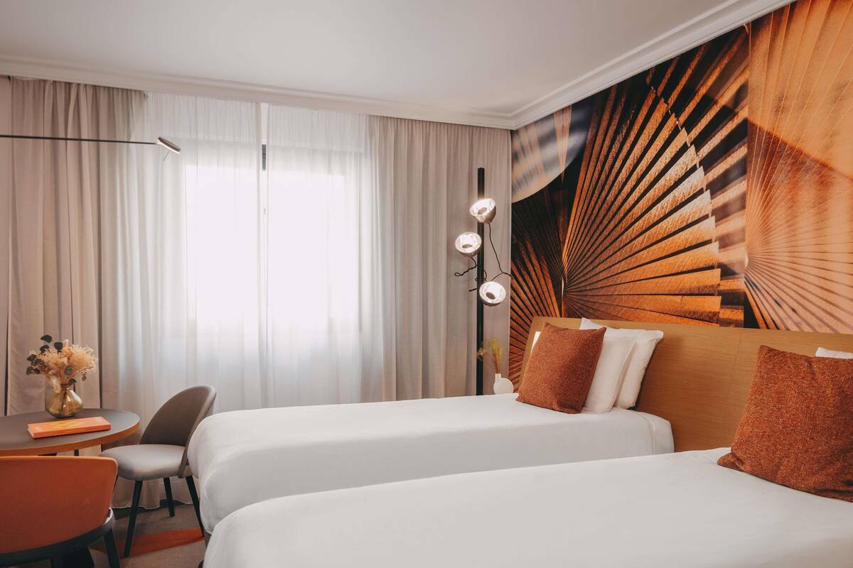 A hotel room with two beds, a desk, a chair, and a large window with curtains.