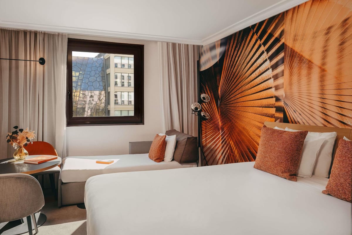 Hotellihuone, jossa on kaksi sänkyä, työpöytä, tuoli, ikkuna verhoilla ja näkymä rakennuksiin.