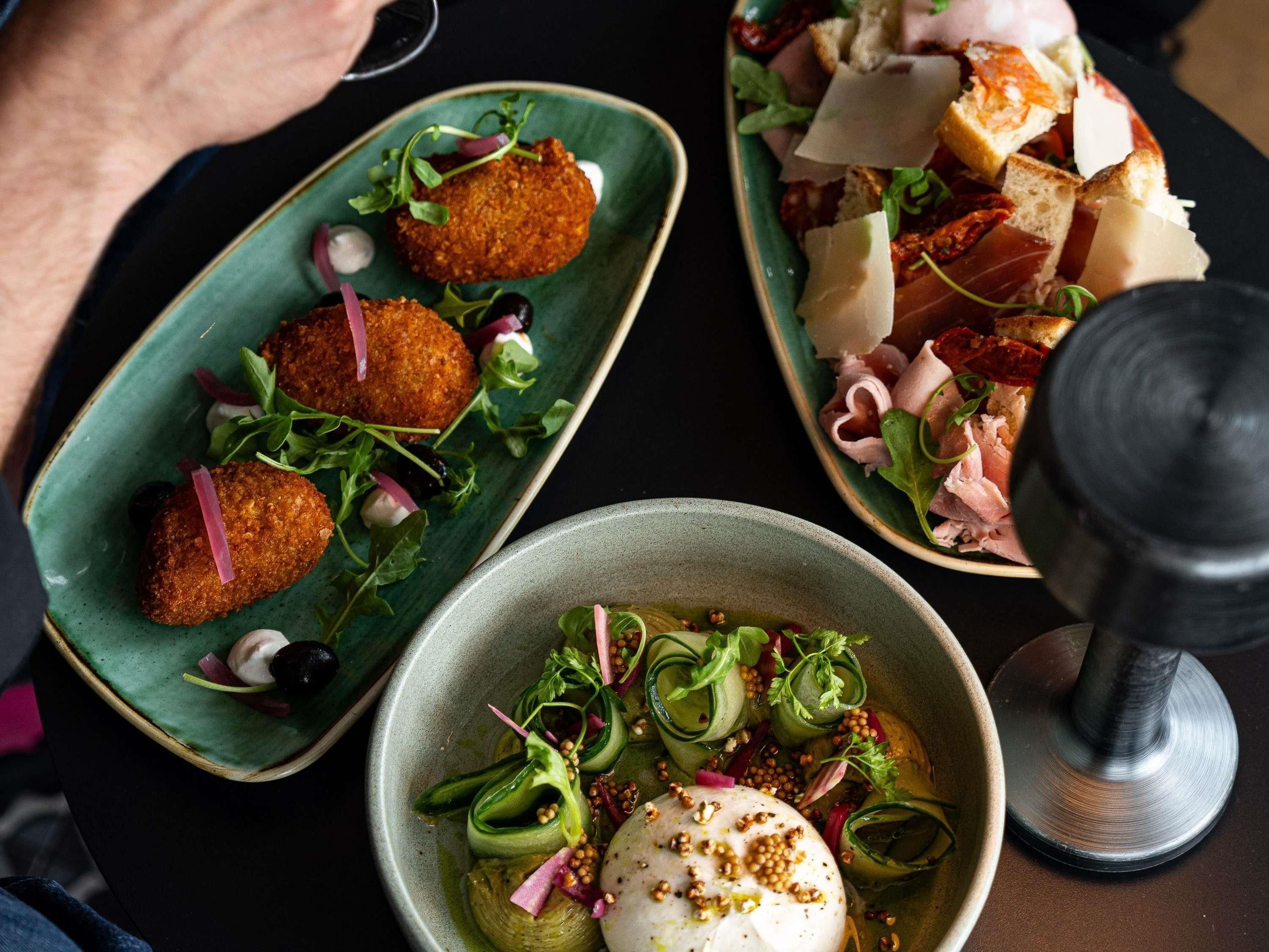 Une table avec trois plats : deux assiettes de mets panés accompagnés de légumes verts, un bol contenant un œuf poché et des légumes verts, et une assiette avec de la viande et du fromage.