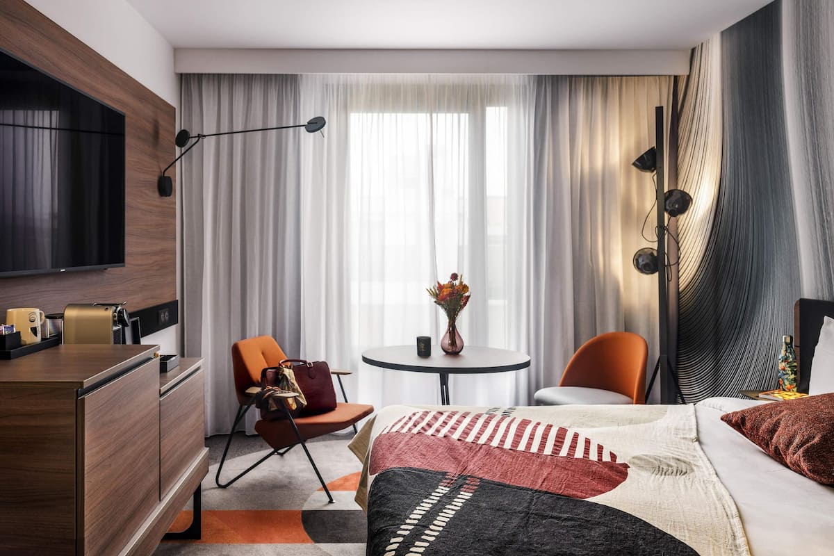 A hotel room with a bed, a desk, a chair, a small table, and a TV.