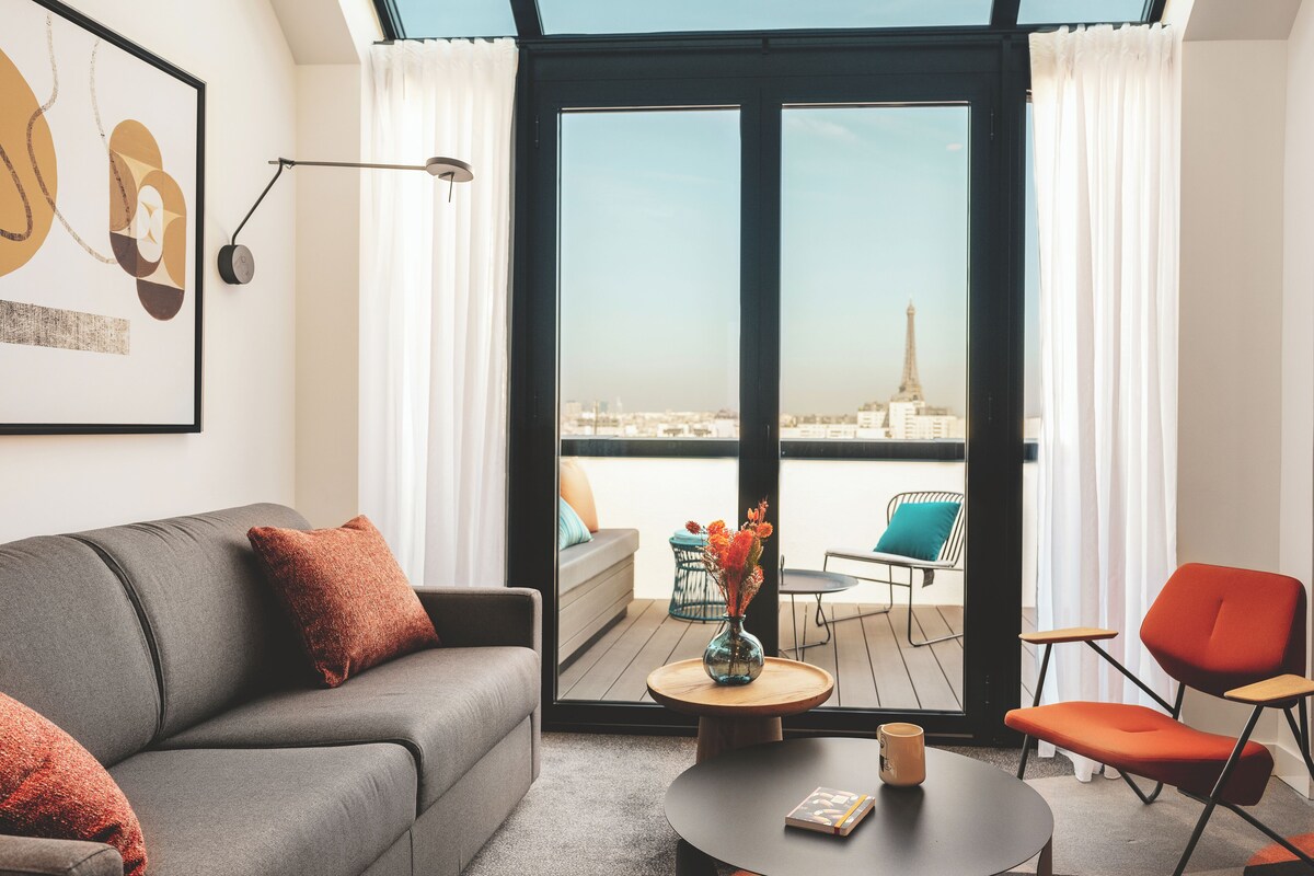 A modern living room with a gray sofa, a red chair, a small round table, and a view of the Eiffel Tower through large sliding doors.