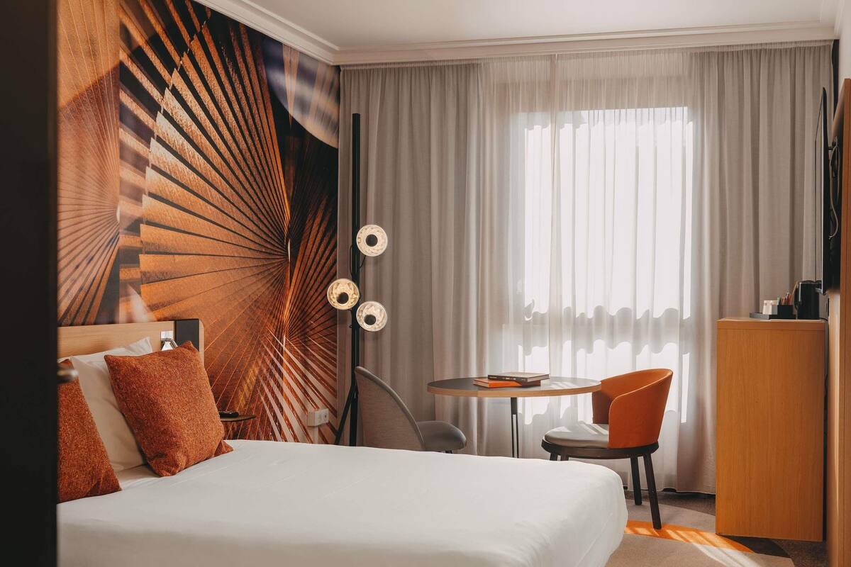 A hotel room with a bed, a desk, a chair, a wardrobe, and a large window with curtains.
