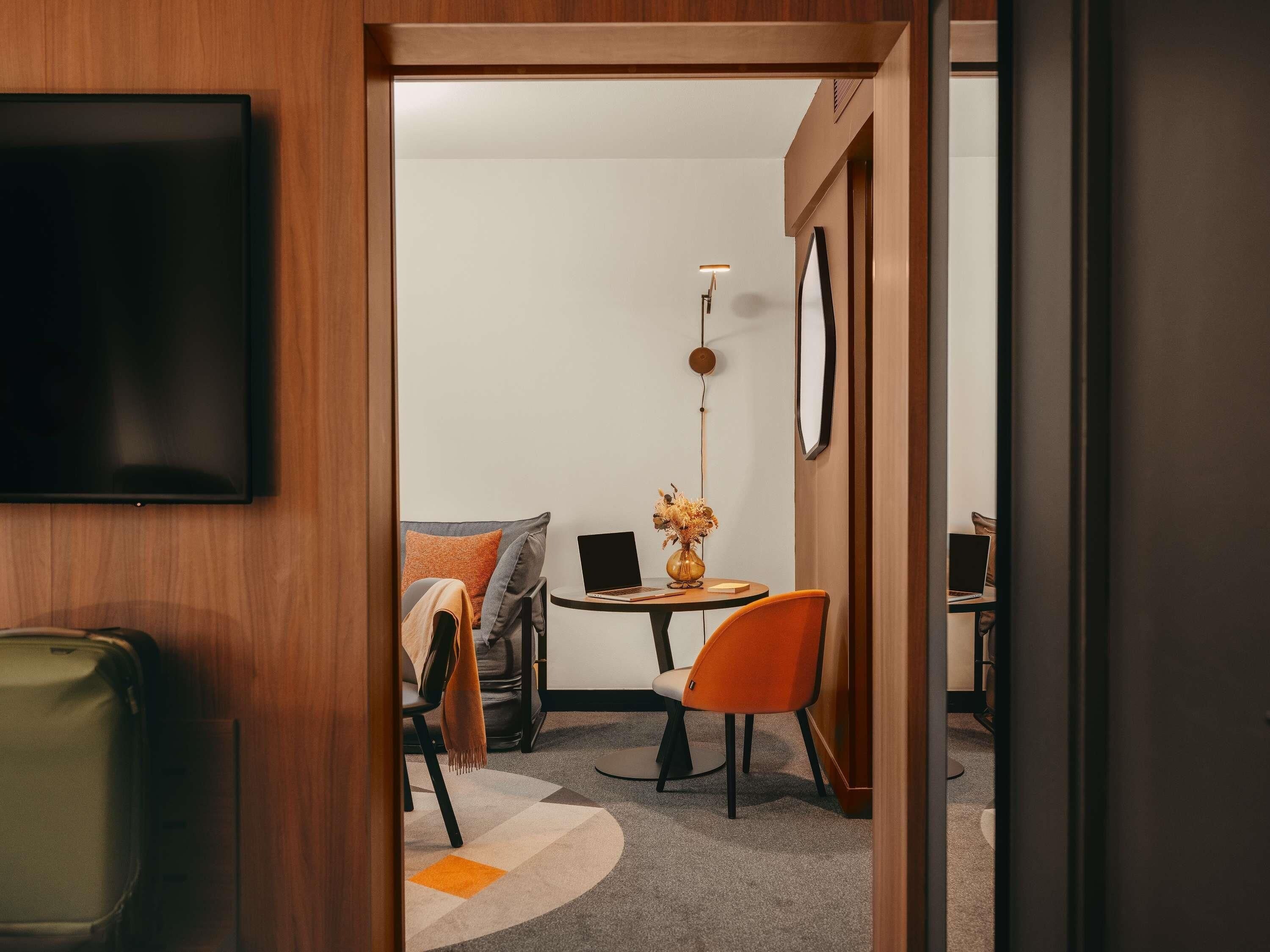 Une chambre d’hôtel équipée d’une télévision, d’un bureau avec un ordinateur portable, d’une chaise, d’une petite table avec un vase et d’une lampe.