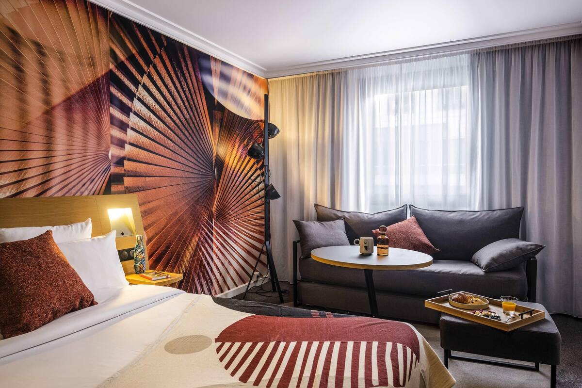 A hotel room with a bed, a sofa, a small table with a tray of breakfast, and a large window with curtains.