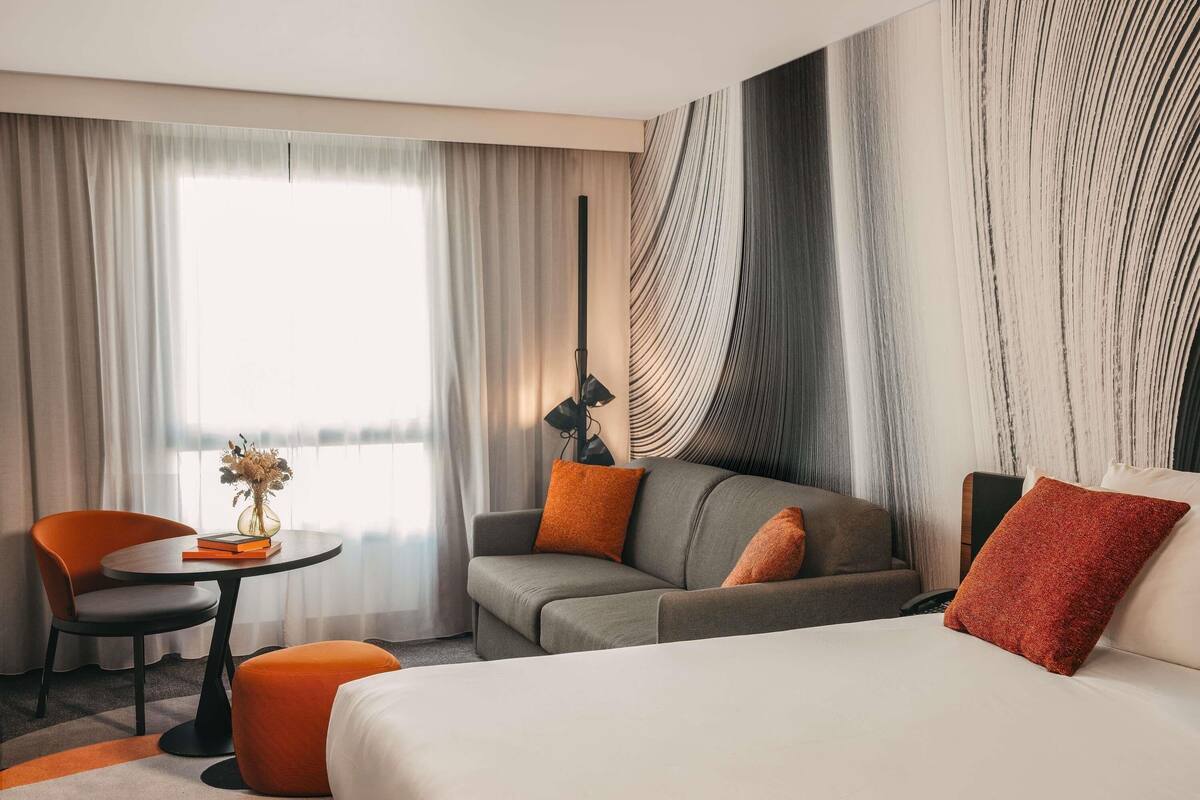 A hotel room with a bed, a sofa, a chair, a small table, and a lamp.