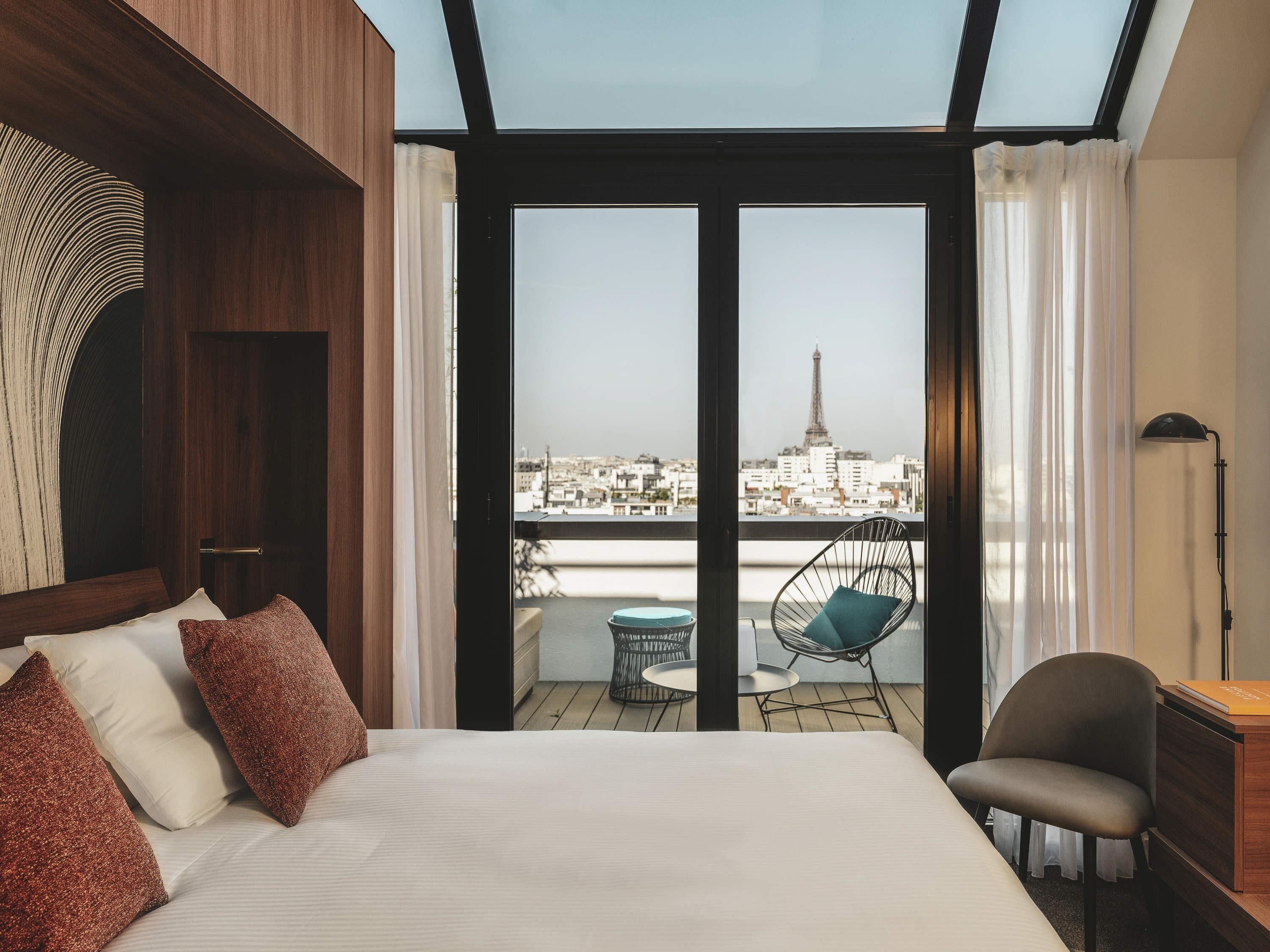 Habitación de hotel con cama, una silla, una mesa y vista a un paisaje urbano que incluye una torre.