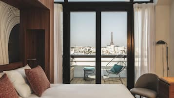 A hotel room with a bed, a chair, a table, and a view of a cityscape including a tower.