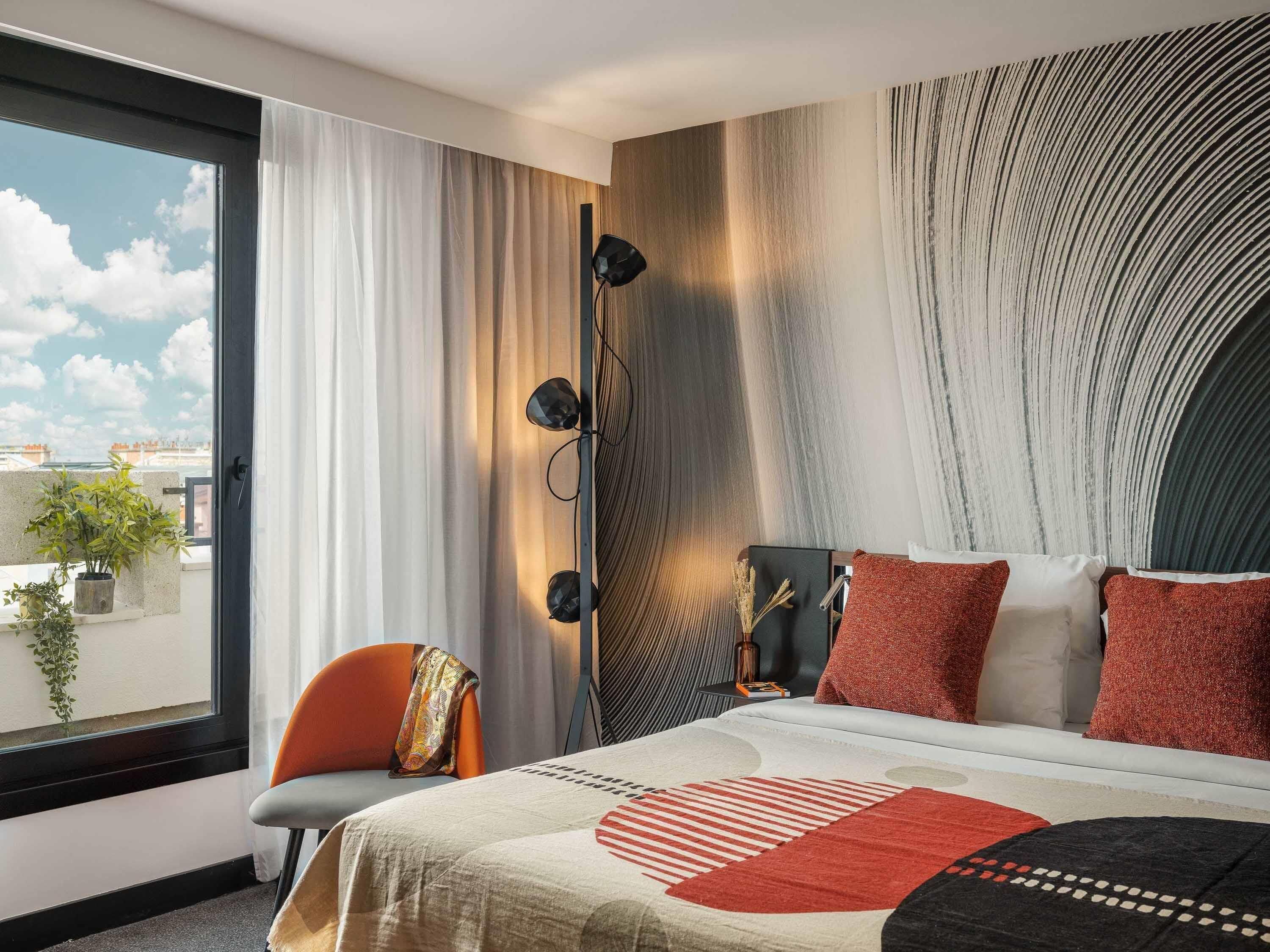 A modern hotel room with a large bed, a view of the city, and a balcony with plants.