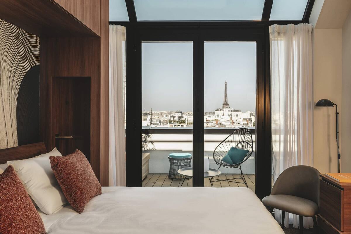 A hotel room with a bed, a chair, a table, and a view of a cityscape including a tower.