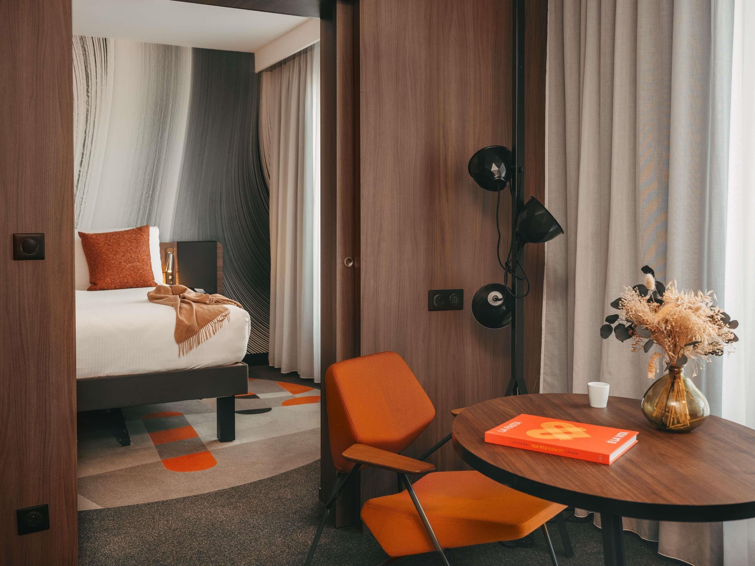 A hotel room with a bed, a desk with a chair, a table with a vase and book, and a lamp.
