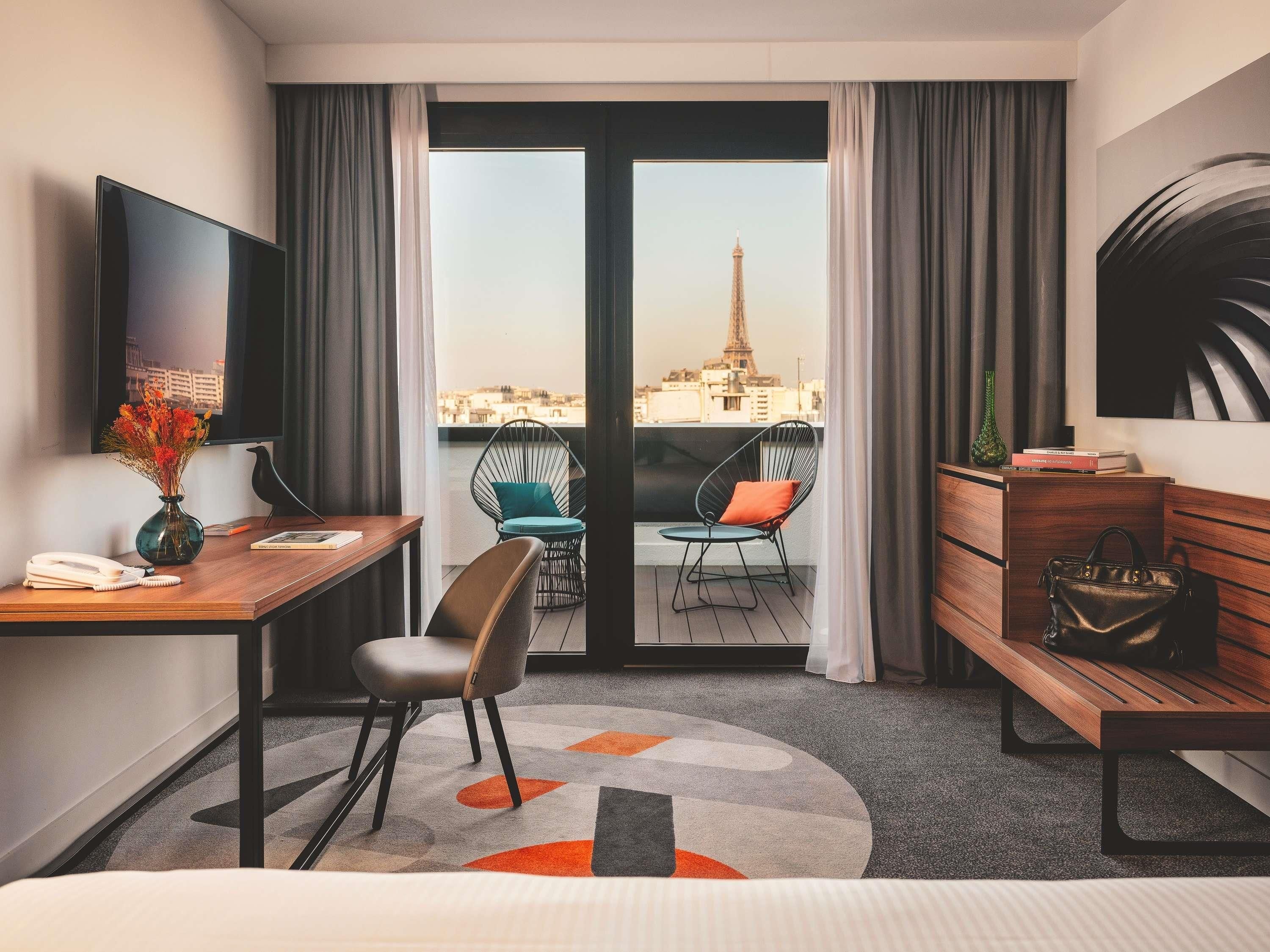 A hotel room with a desk, chair, bed, and a view of the Eiffel Tower.